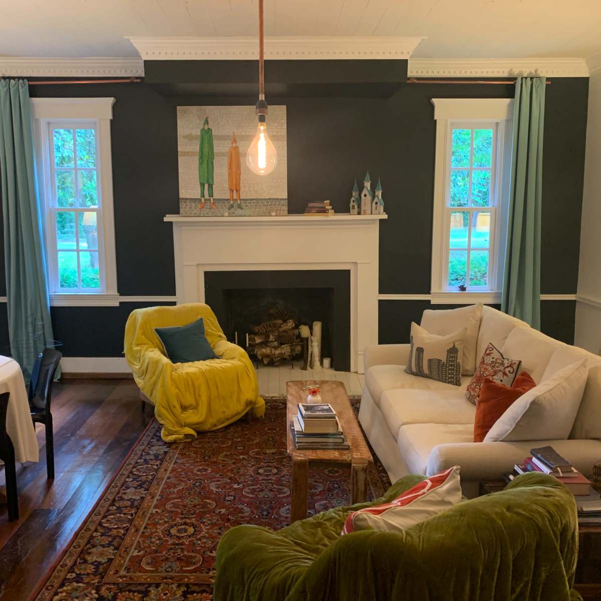 A cozy living room features a white sofa, colorful chairs, a fireplace, and large windows with turquoise curtains.