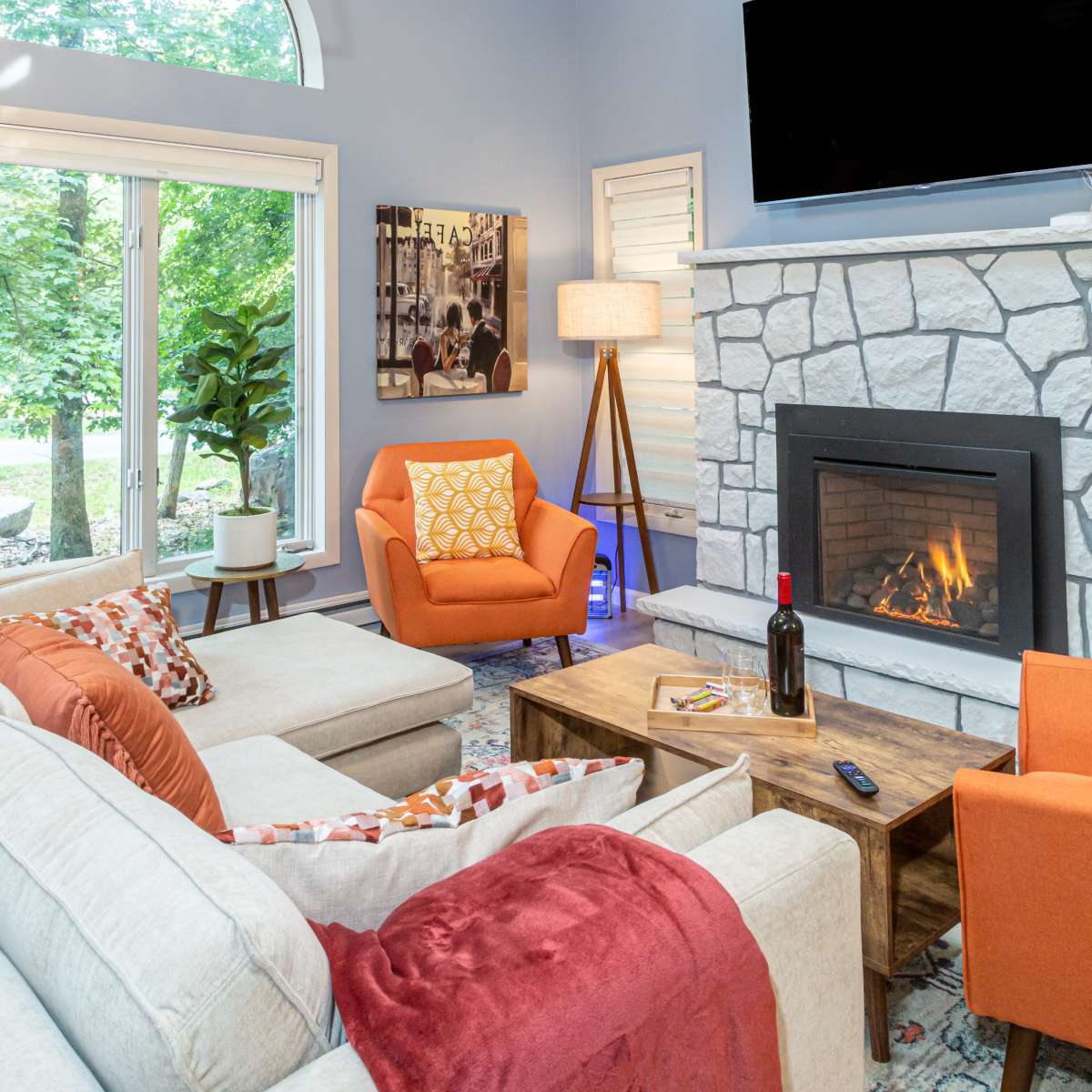 The living room features a stone fireplace, a wooden coffee table, and an orange armchair, with large windows allowing natural light to fill the space.