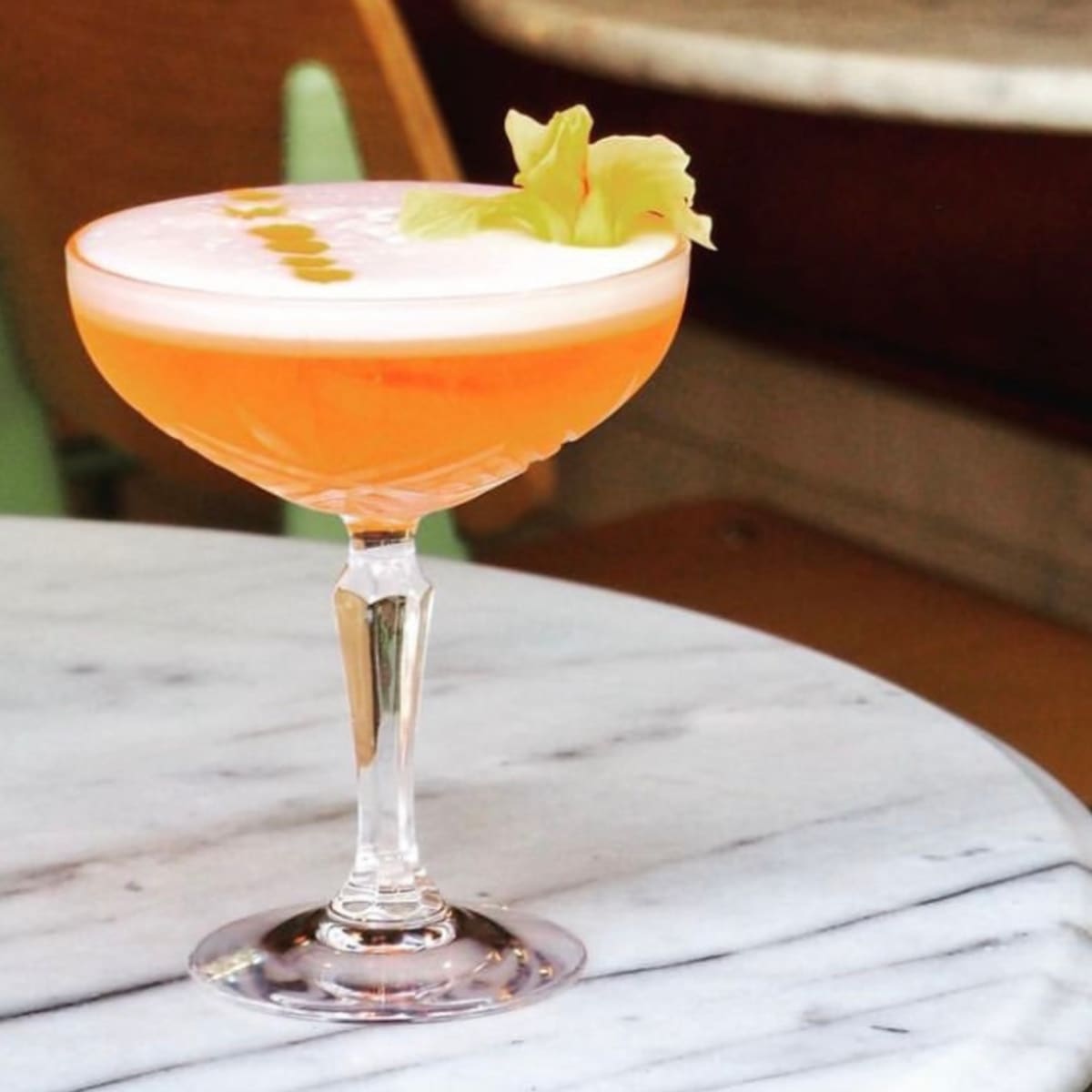 A cocktail in a glass garnished with a flower sits on a marble table next to wooden chairs.