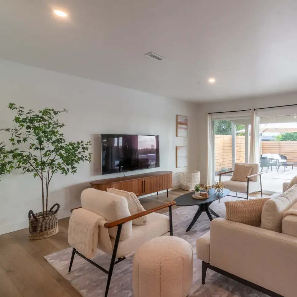 A modern living room features a white sofa set, a wooden TV console, and large sliding glass doors that open to an outdoor patio area.
