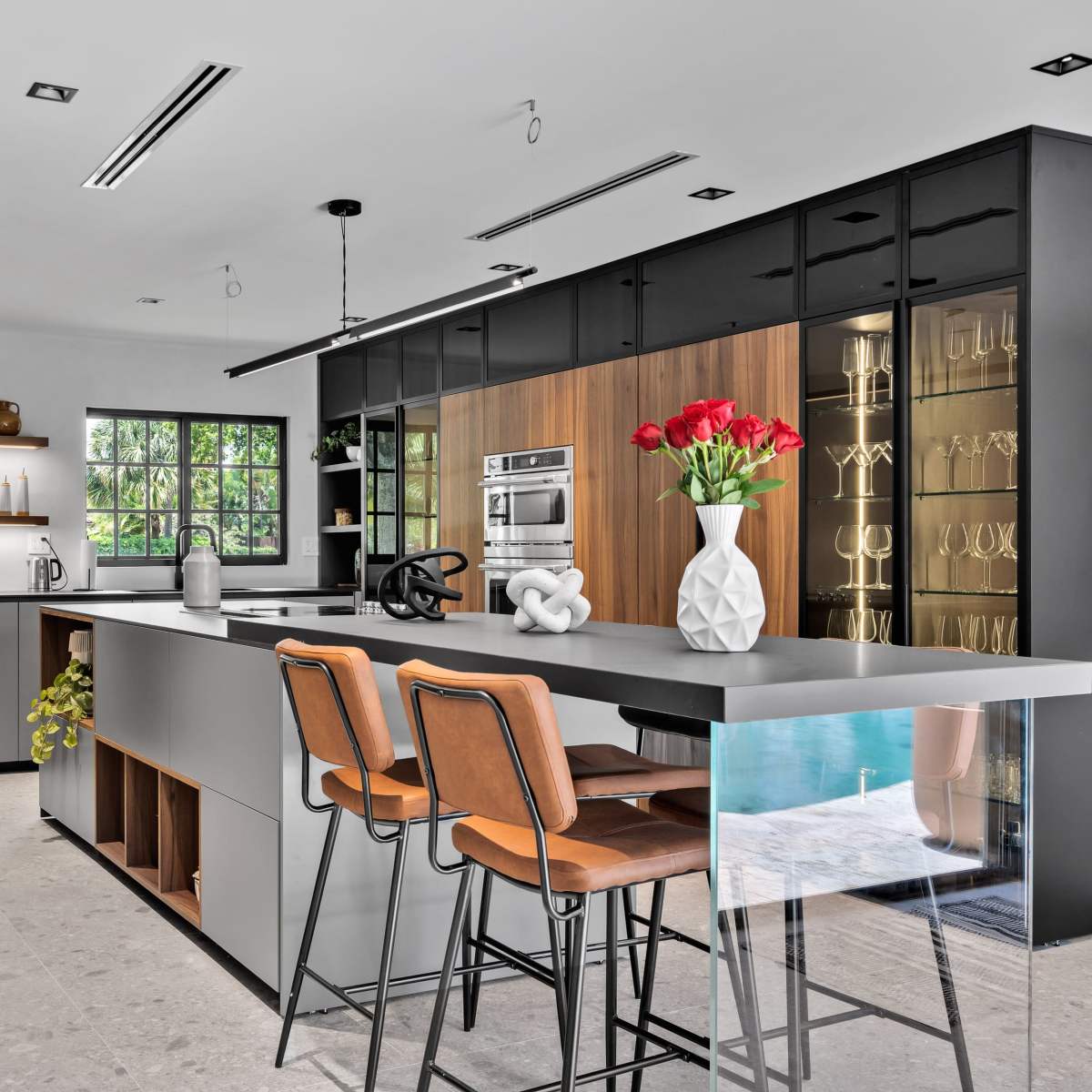 A modern kitchen features sleek cabinetry, an island with bar stools, and large windows providing natural light.