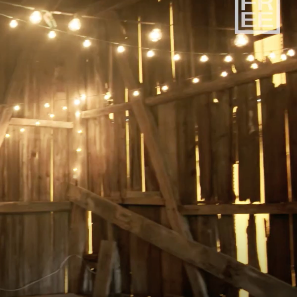 The interior of a wooden barn features string lights illuminating the space and various pieces of wood and furniture stored along the walls.
