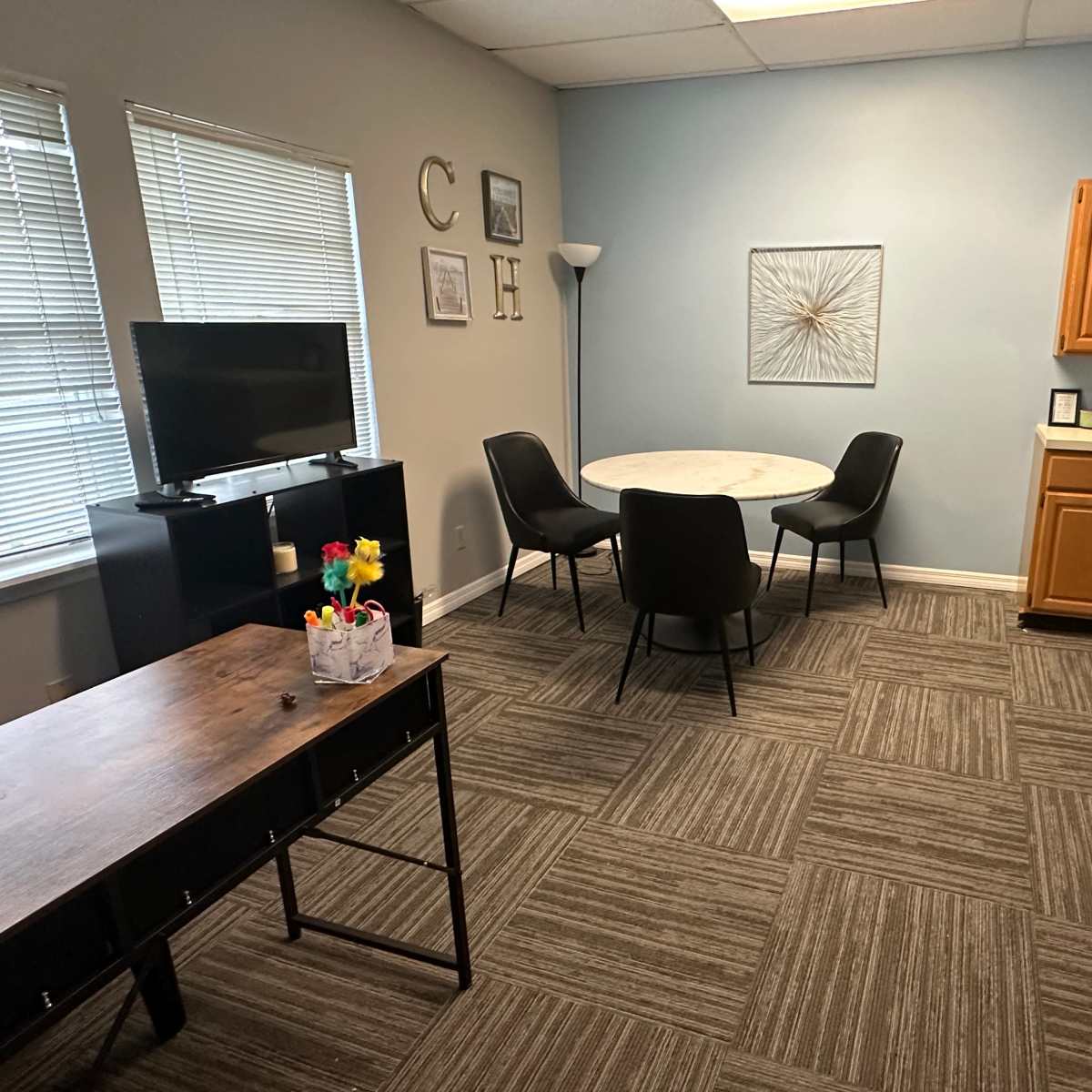 The room features a small dining area with a round table and four chairs, a television, a desk with office supplies, and a kitchenette with wooden cabinets.