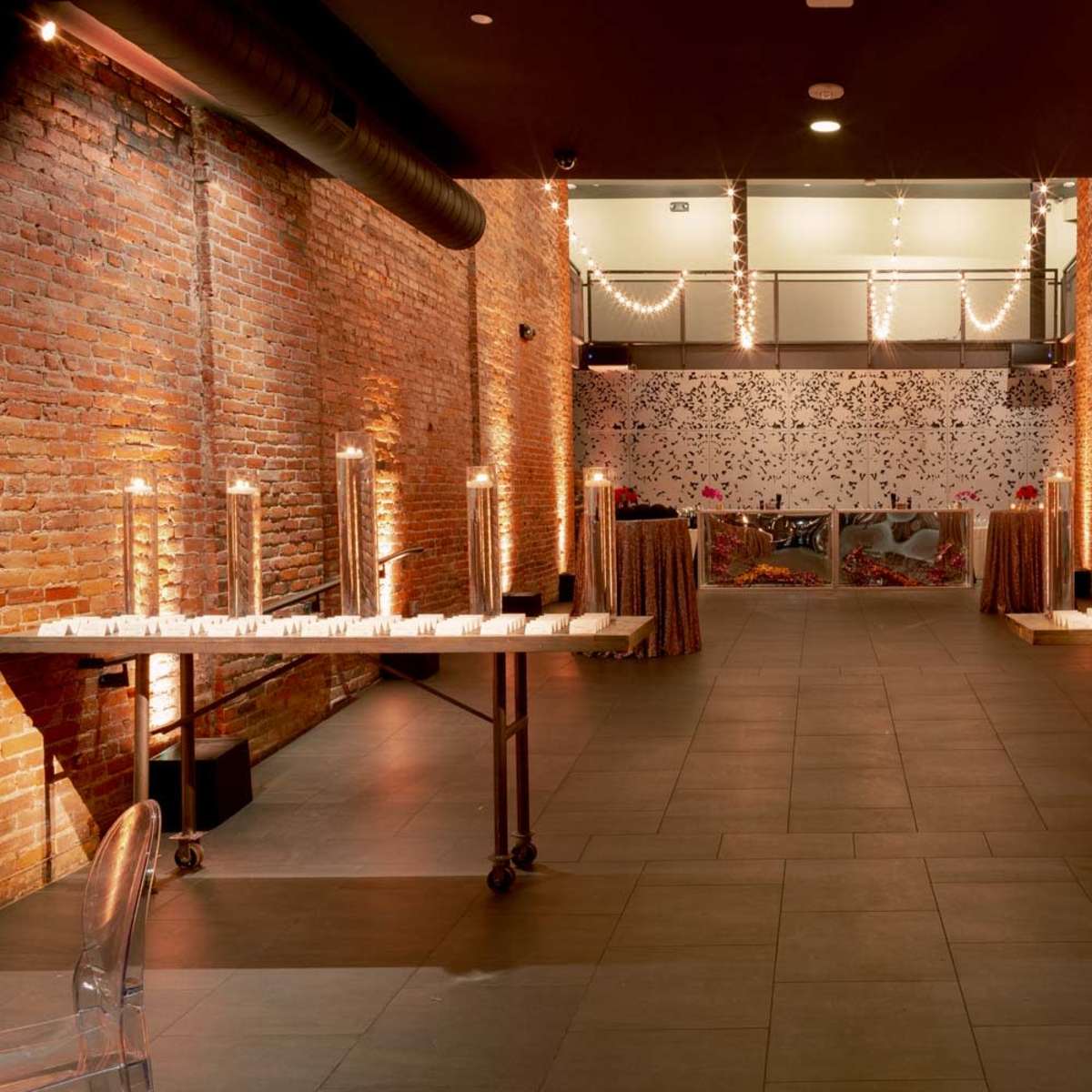 A spacious event venue with exposed brick walls, two tables arranged on either side adorned with candles, and decorative elements at the back.