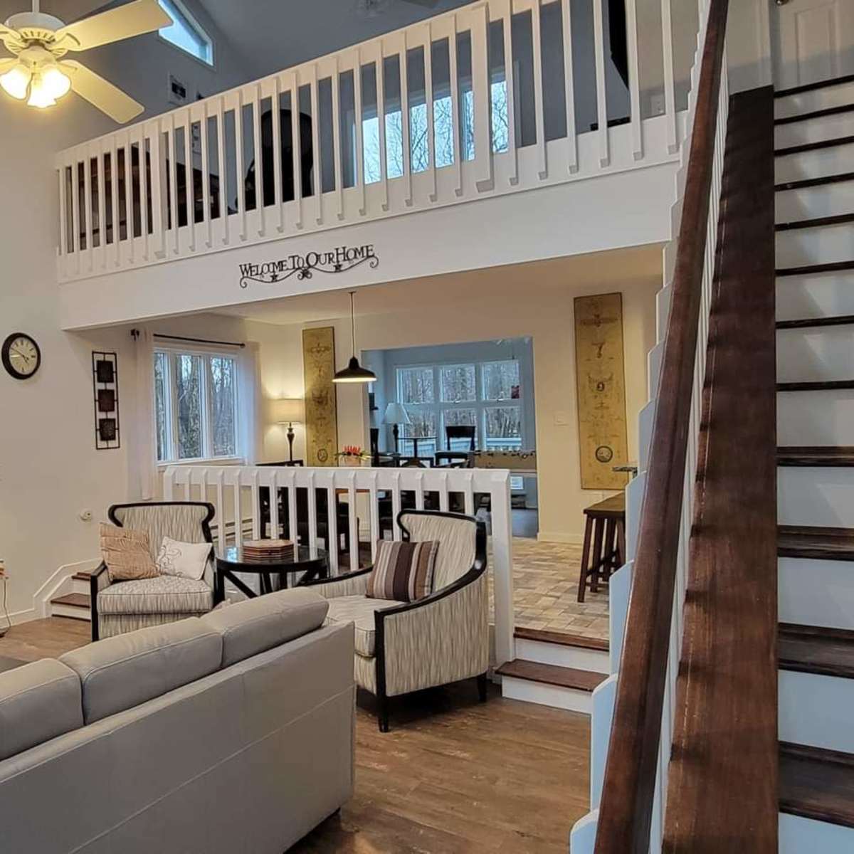 A spacious living room with a stone fireplace, large windows, and a staircase leading to a loft area.