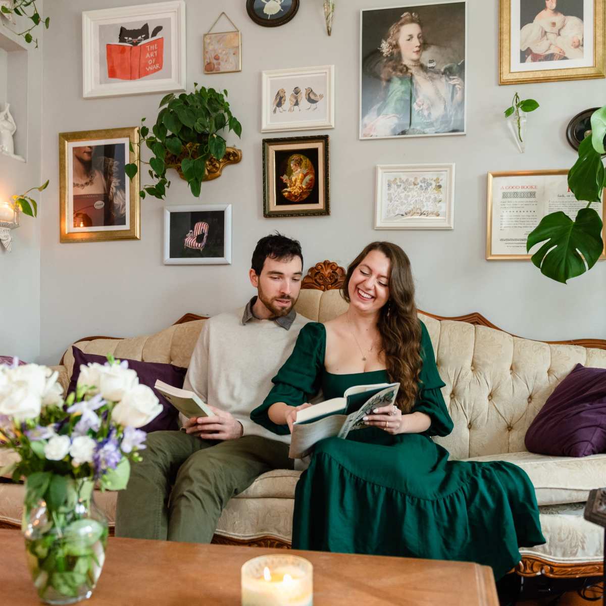 A couple sits together on a vintage sofa in a cozy living room filled with framed art and plants, reading books.