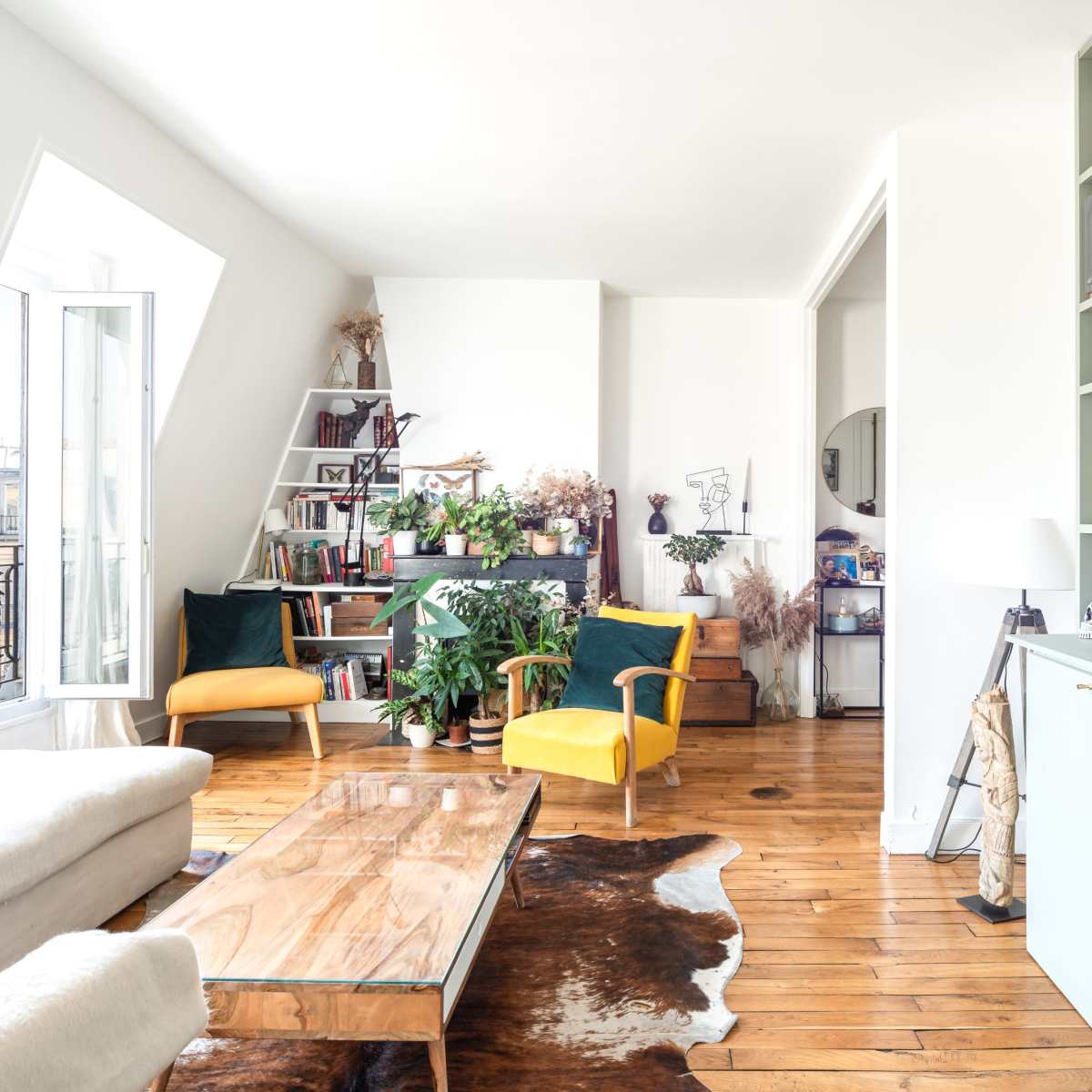 A bright living room features a wooden coffee table, yellow chairs, a bookshelf filled with plants and books, and large windows letting in natural light.