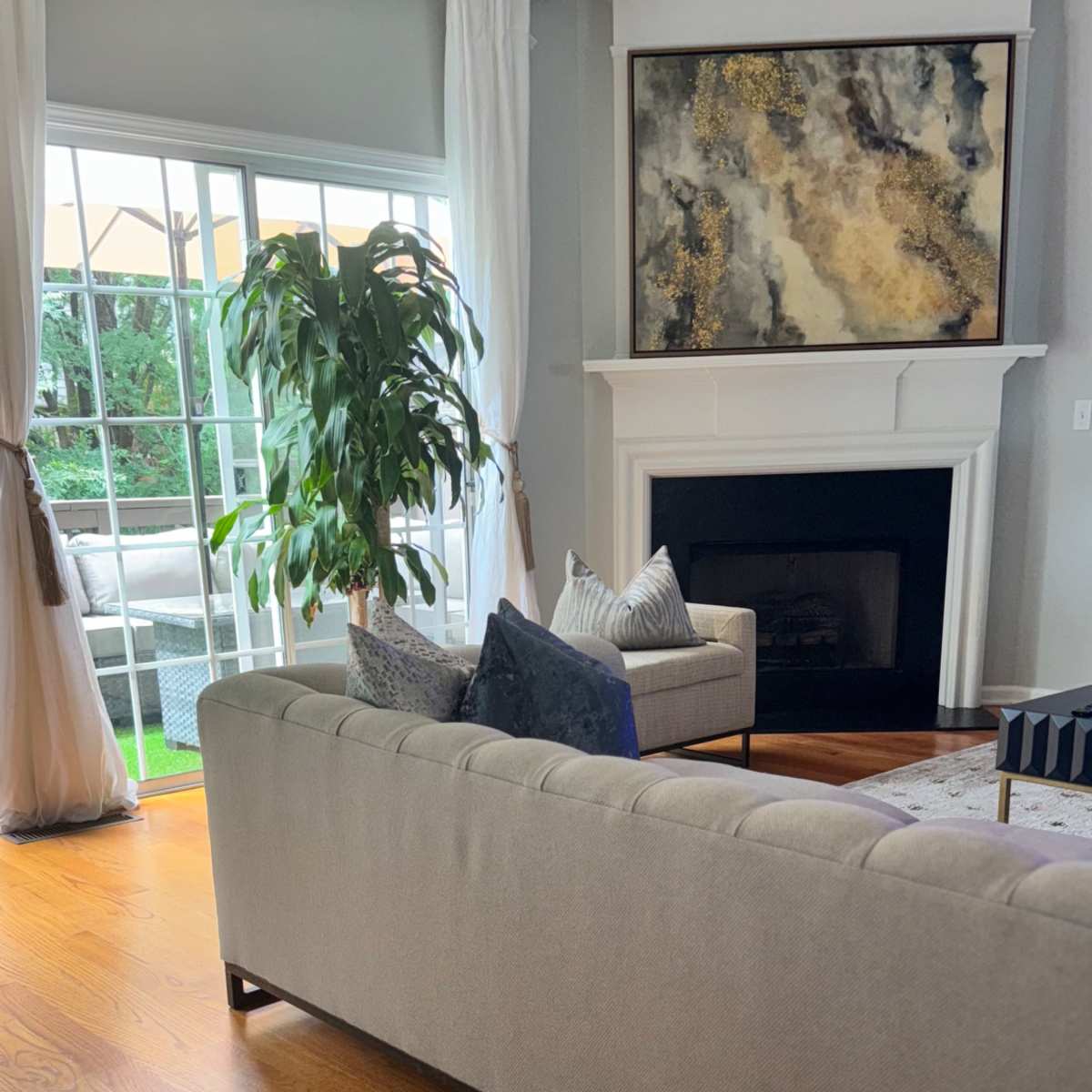 A light-filled living room features a gray sofa, a decorative plant in the corner, and a fireplace beneath a large piece of wall art.