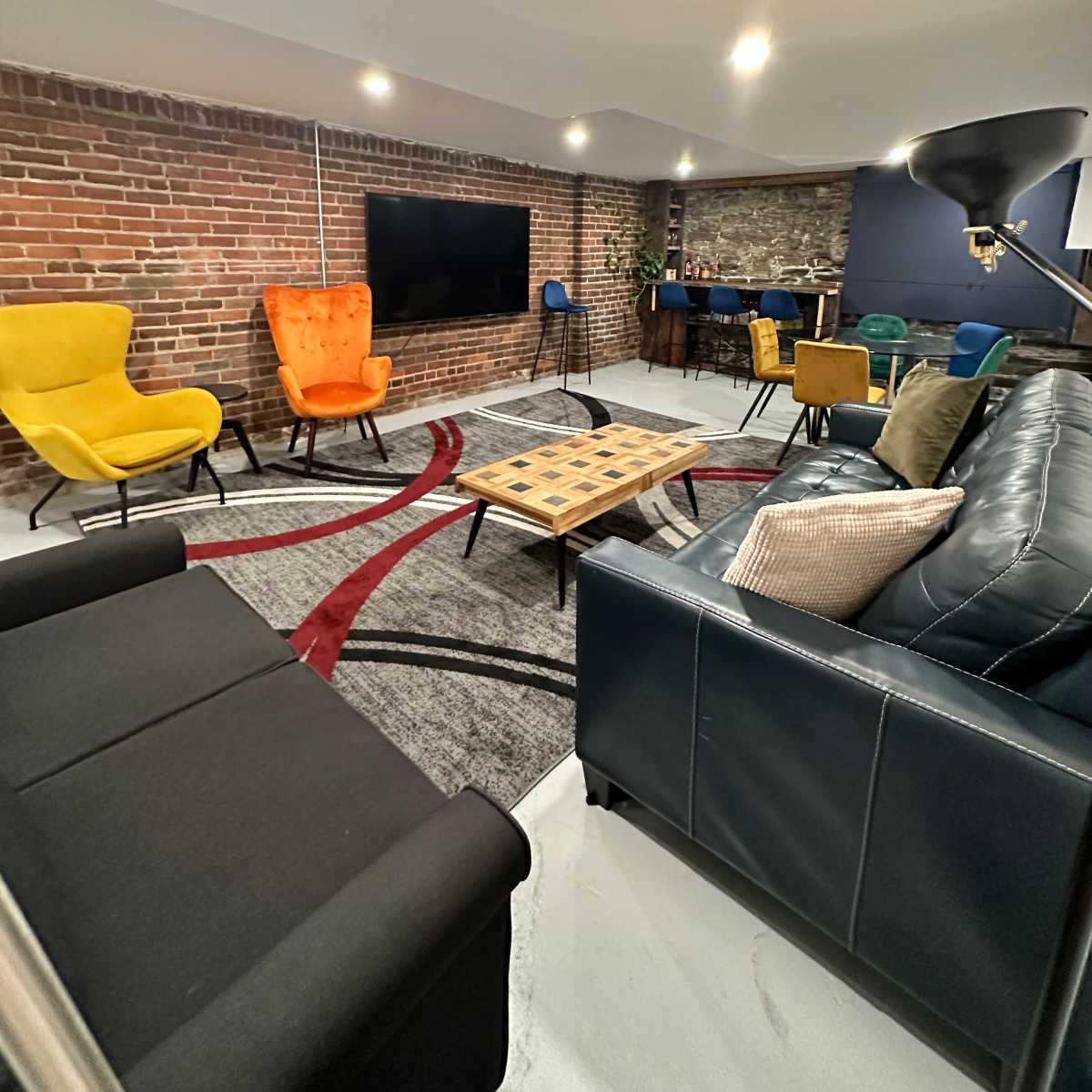 A modern lounge area featuring a mix of colorful chairs, a coffee table, and a large television against a brick wall.
