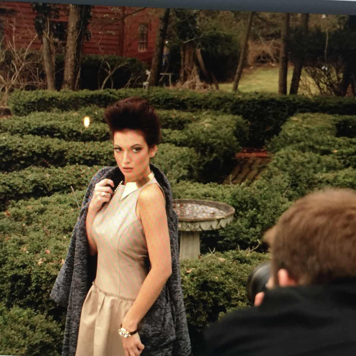 A model poses in a garden with neatly trimmed hedges while a photographer captures her image.
