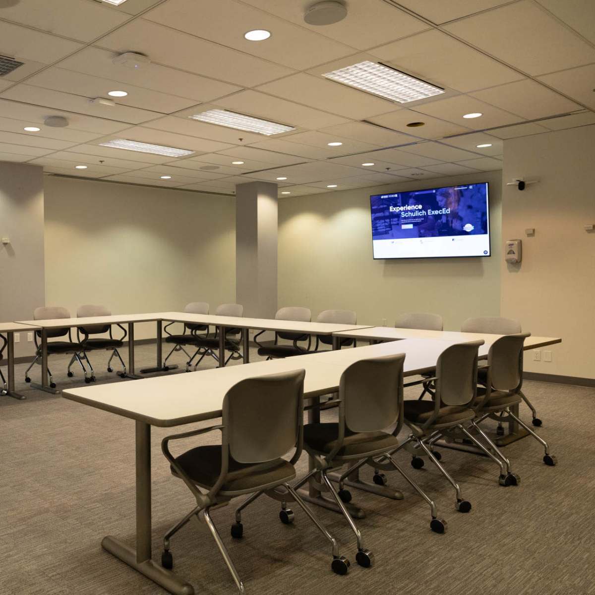 A modern conference room features a large table surrounded by chairs, a wall-mounted screen, and windows providing natural light.