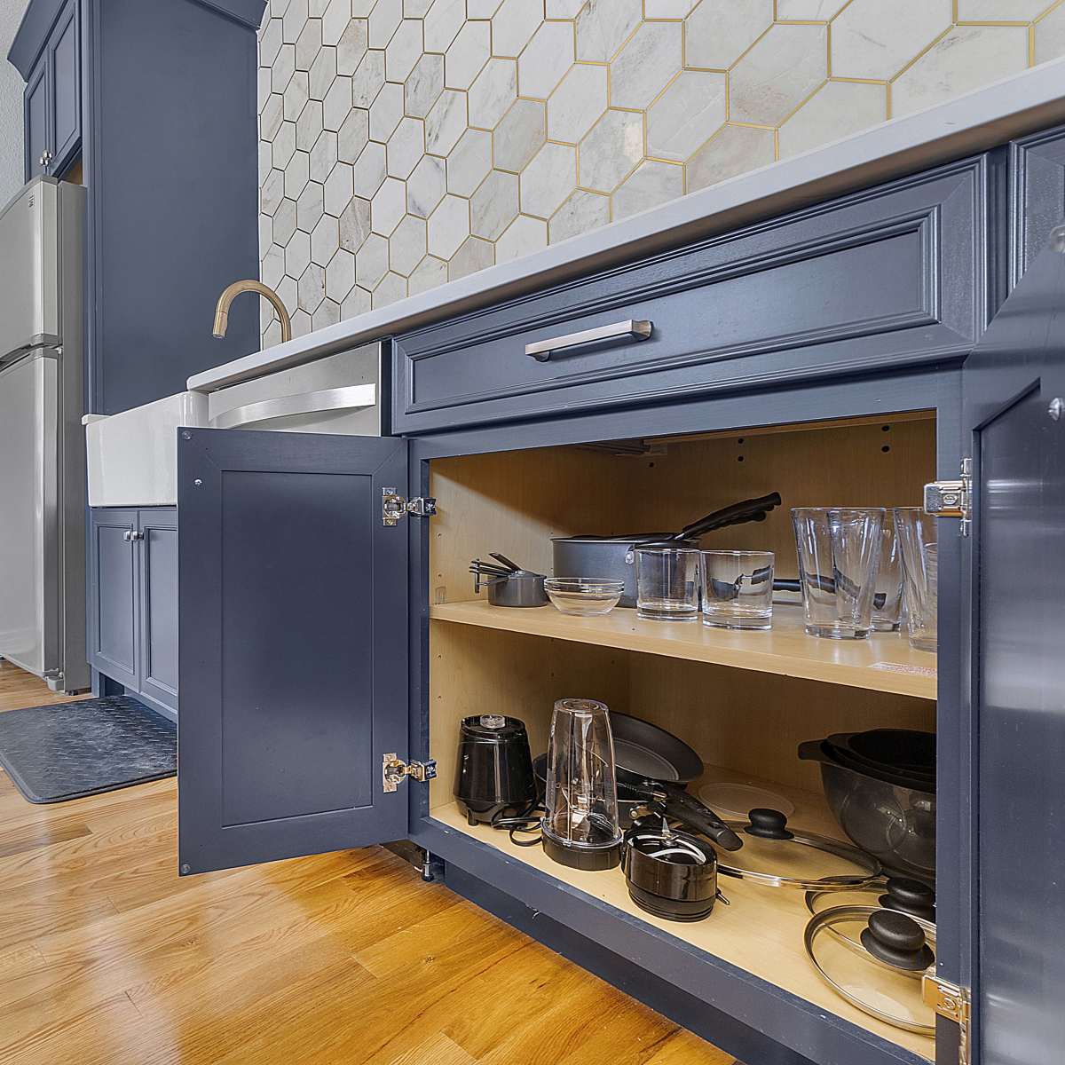 A kitchen with navy blue cabinets and a hexagonal tile backsplash, featuring an open cabinet displaying various kitchen utensils and glassware.