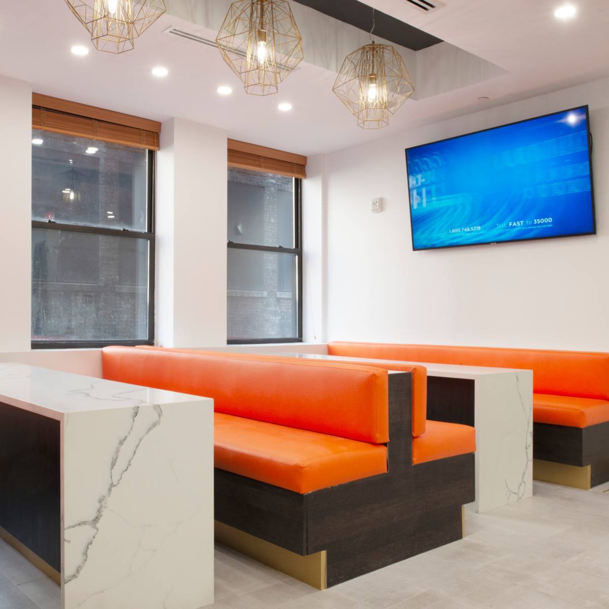 A modern, minimalist dining area featuring orange upholstered benches, a marble-topped table, and large windows with wooden blinds.