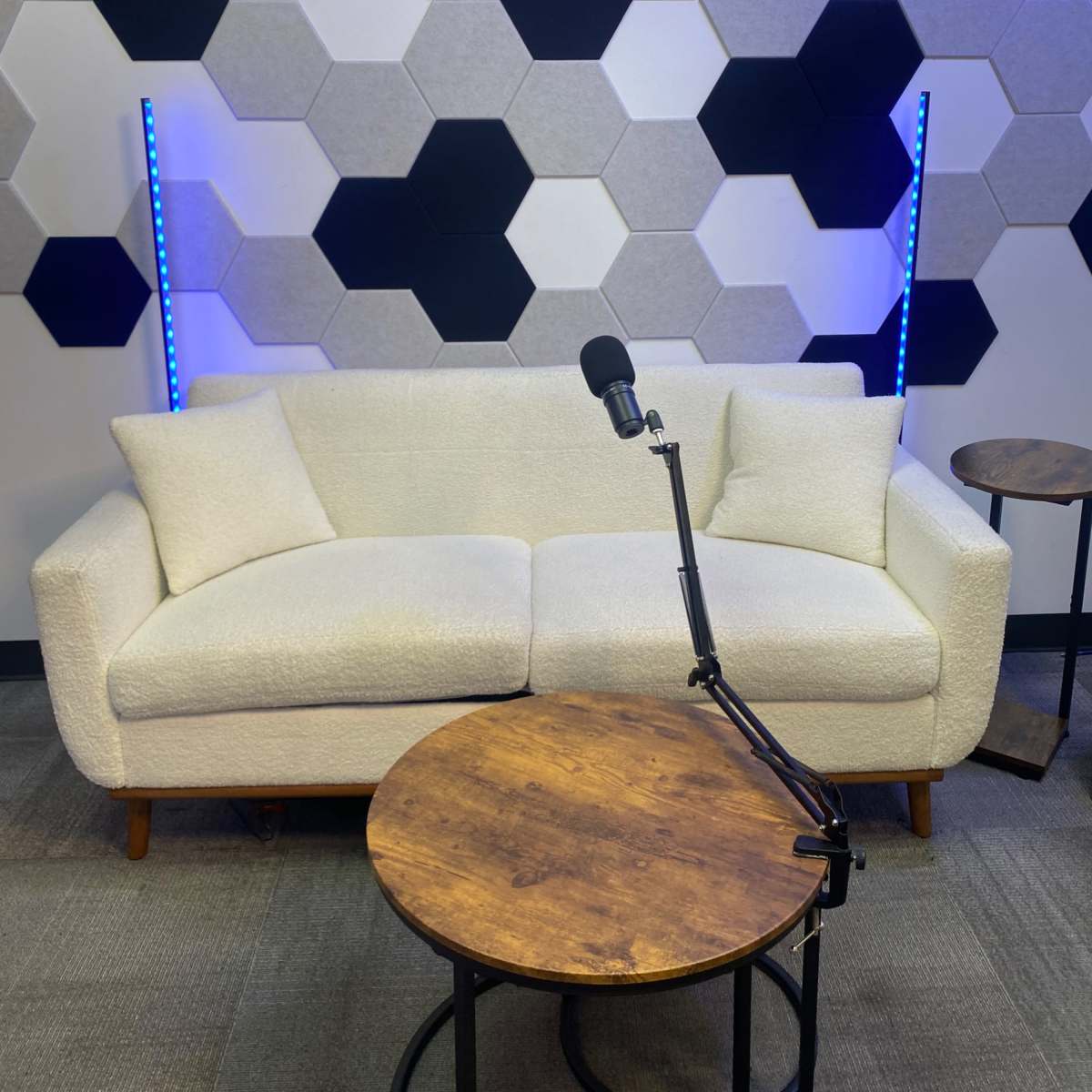 A light-colored couch is positioned in front of a hexagonal-patterned wall, with a microphone on a stand and two small wooden tables nearby.