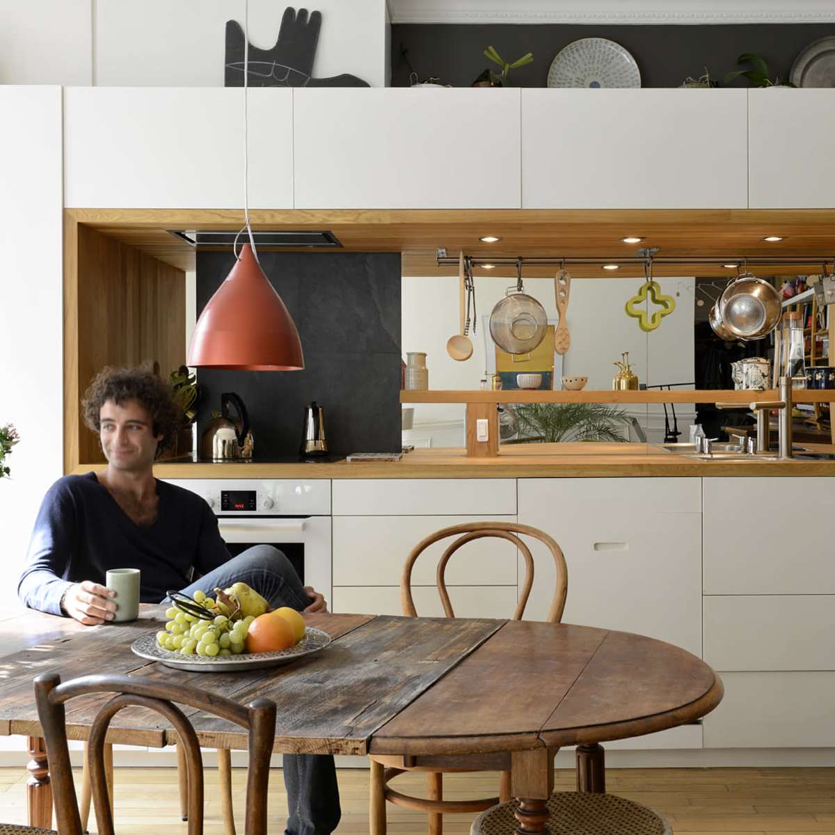 A man sits at a wooden dining table with a fruit bowl in front of him, while an open kitchen with a minimalist design and wooden accents is visible in the background.