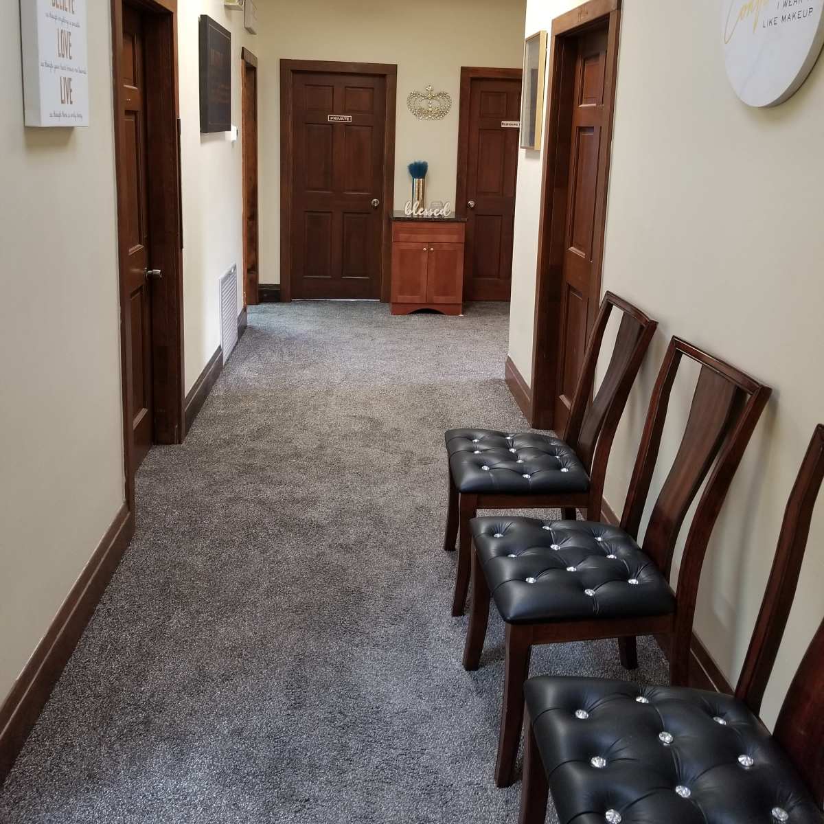 A hallway with light-colored walls, wooden doors on either side, and a row of four wooden chairs positioned against one wall.