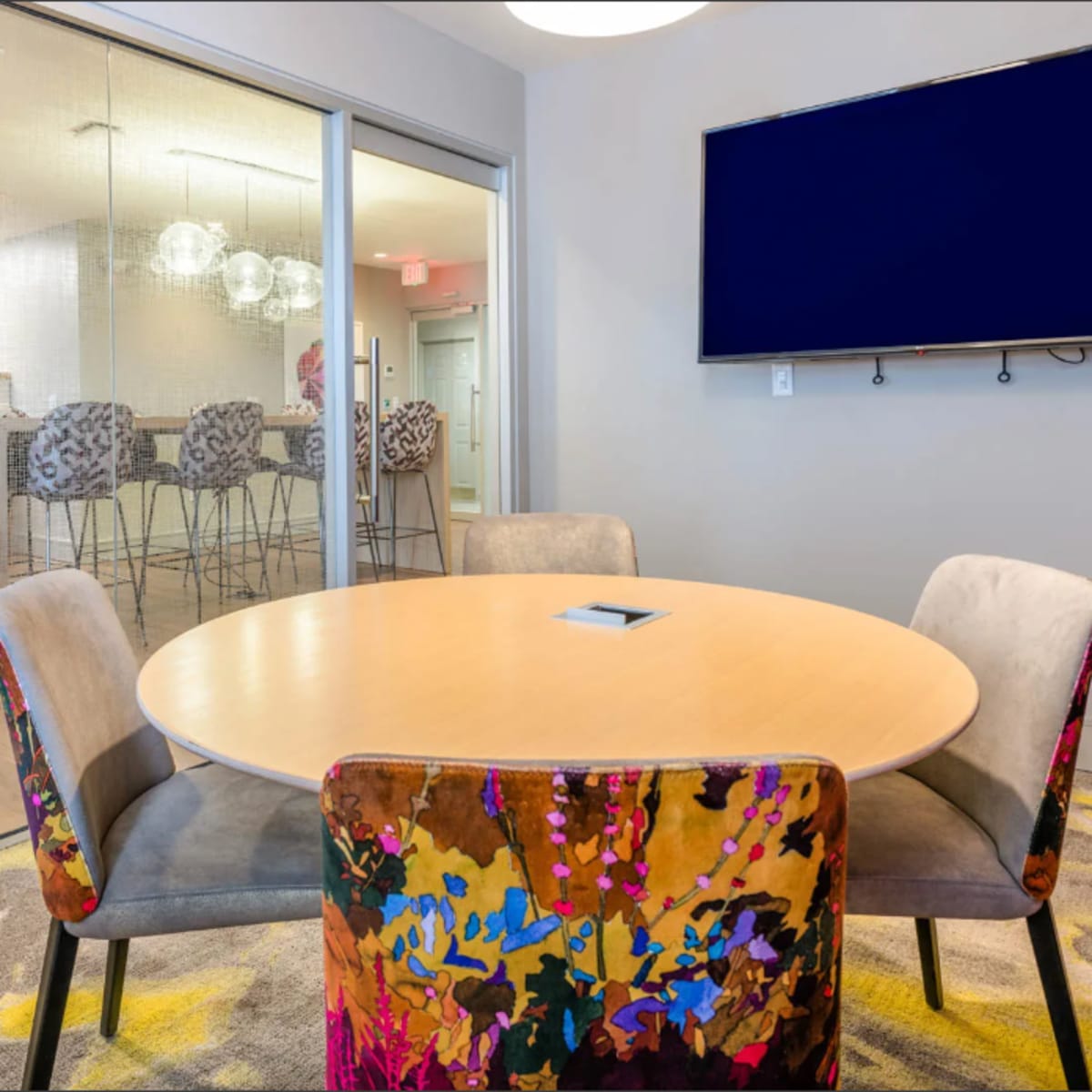 A round table with colorful chairs is set in a modern meeting room, adjacent to a space featuring a kitchen and bar stools, with a large screen mounted on the wall.