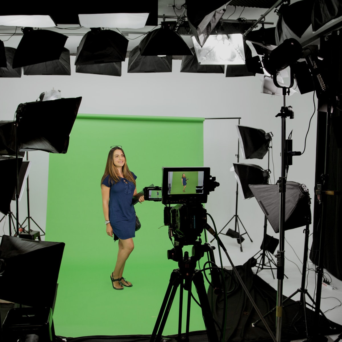 A woman stands on a green screen set surrounded by studio lights and camera equipment.