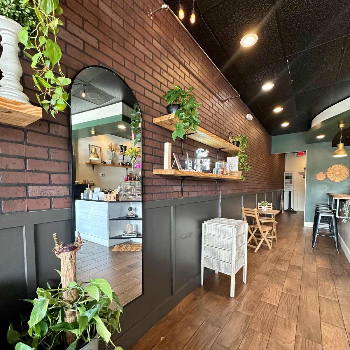 A cozy café interior with brick walls, wooden flooring, and plants along a shelf, leading to a bright service area at the back.