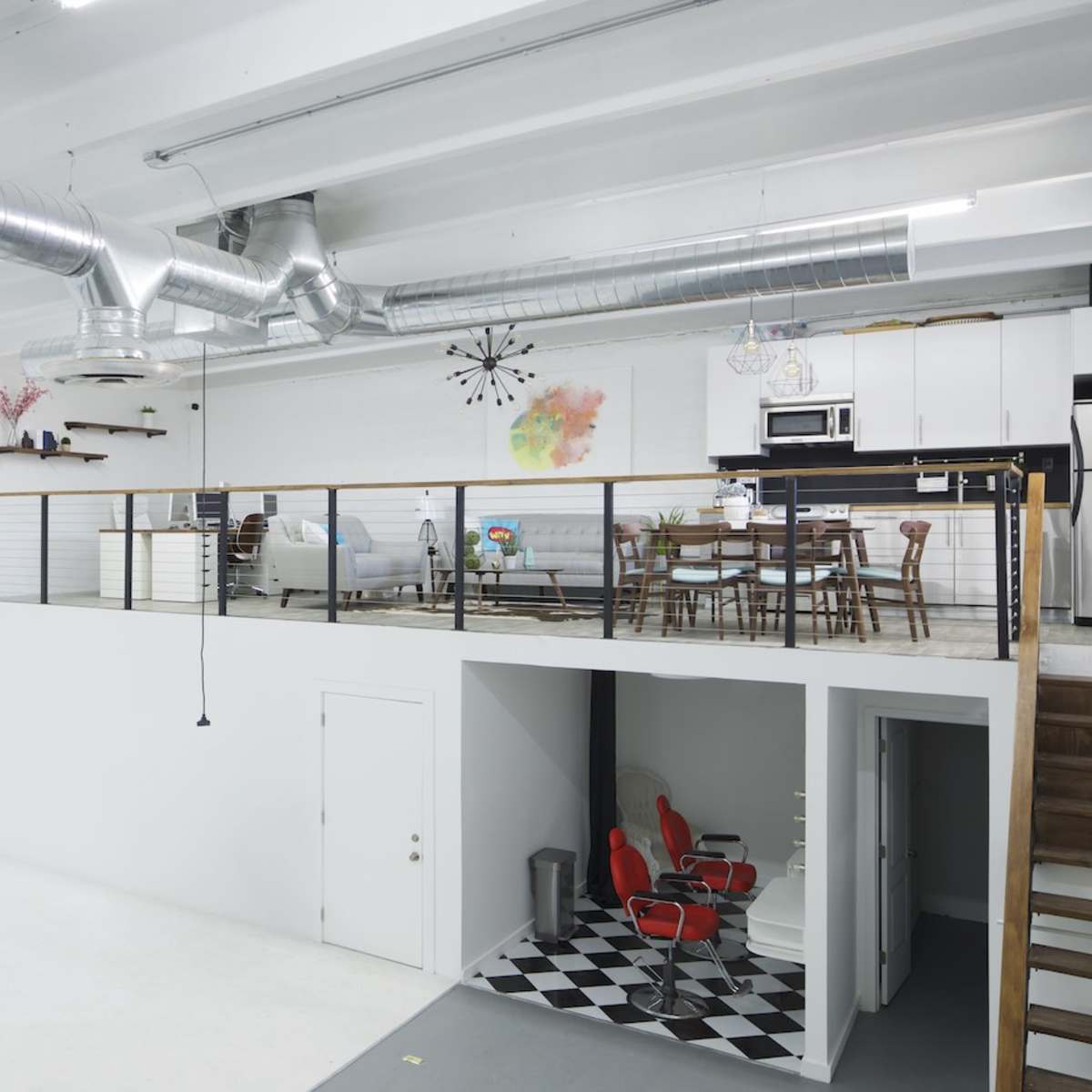 A spacious, modern studio with a loft area, featuring a seating area and dining space, and a lower level with black and white checkered flooring and two red chairs.