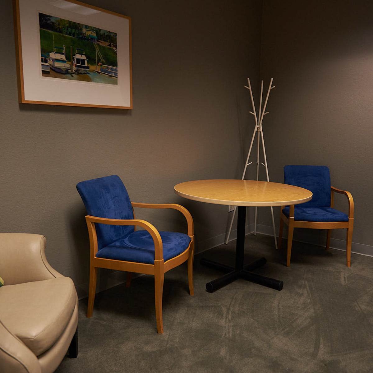 A small, simple waiting area features two blue chairs and a round wooden table beside a beige armchair and a wall-mounted artwork.