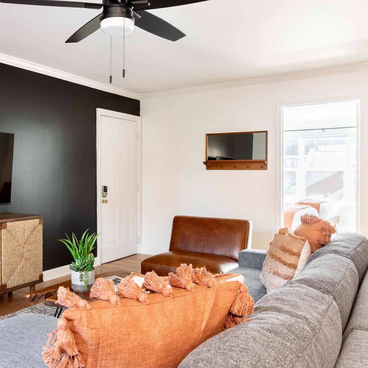 The living room features a black accent wall, a large flat-screen TV on a wooden console, a brown leather chair, and a gray sofa adorned with orange cushions.