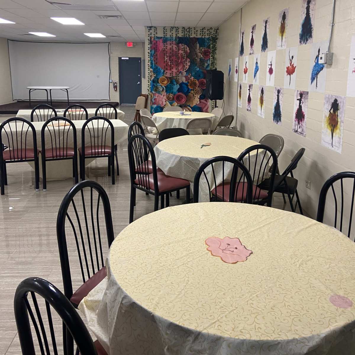 A spacious indoor room set up with round tables covered in light-colored tablecloths, accompanied by chairs and decorated walls featuring various artworks.