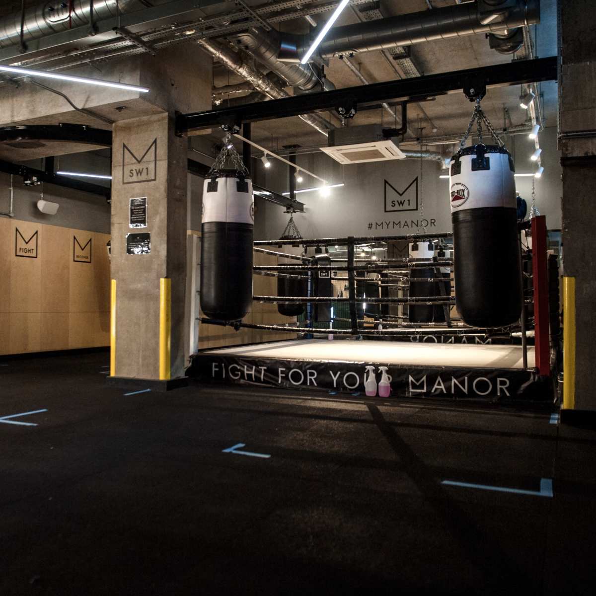 An indoor boxing gym featuring several hanging punching bags and a boxing ring in the background.