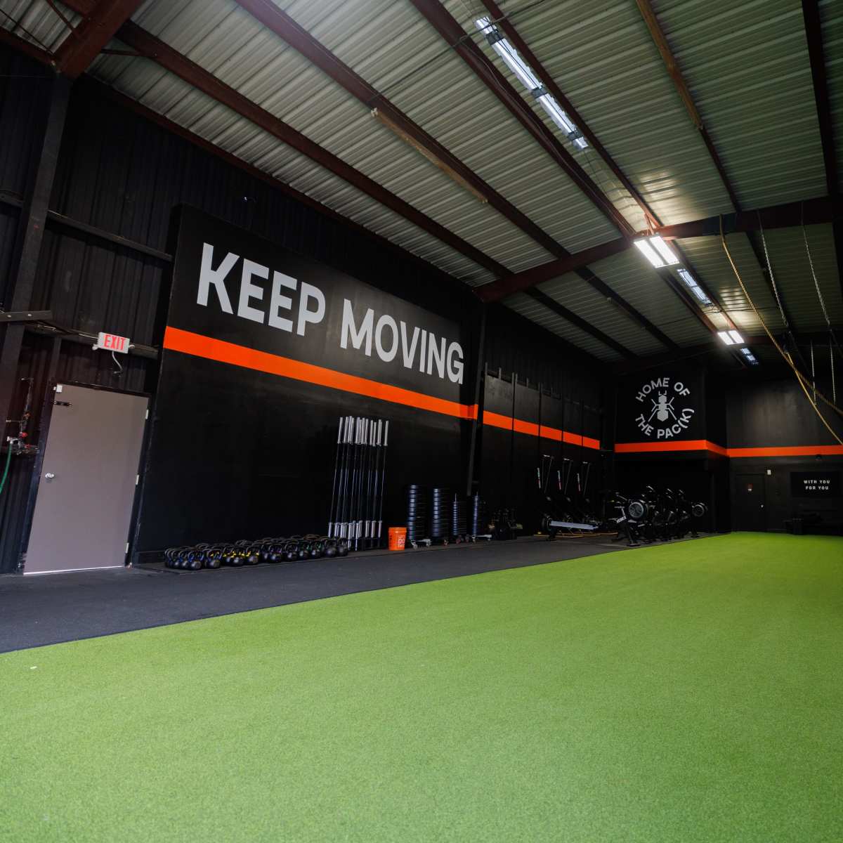 A spacious gym features a green turf floor, black walls with motivational text, and equipment arranged along the sides.