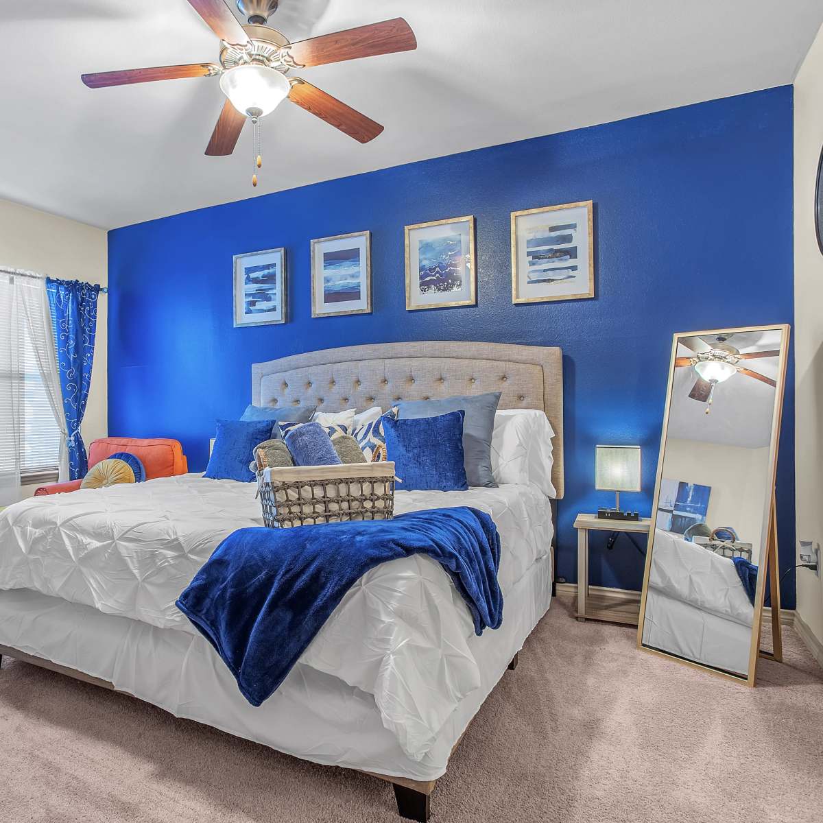A bedroom featuring a large bed with a tufted headboard, blue walls, colorful accents, and a ceiling fan.