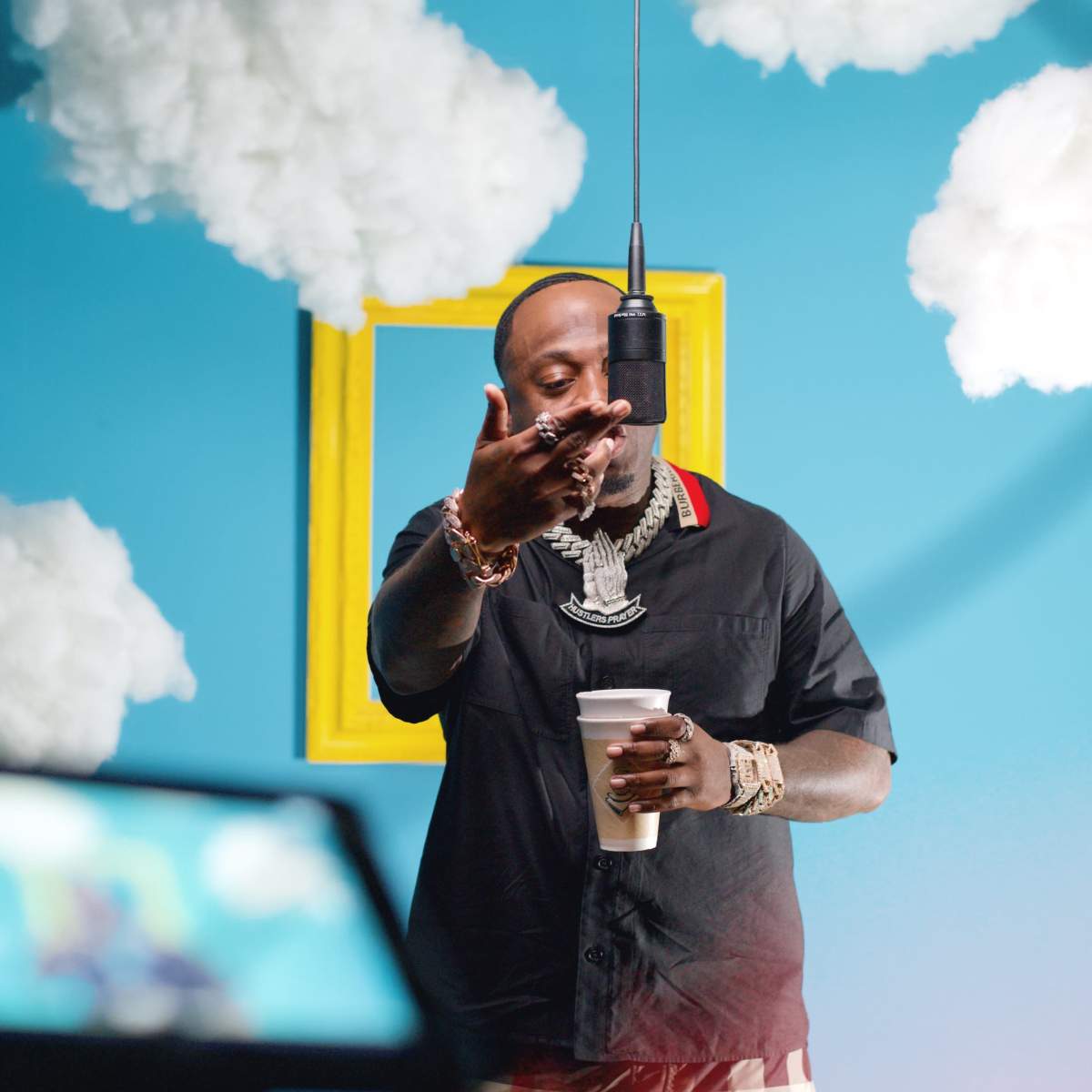 A person holding a cup gestures towards a microphone suspended from above against a bright blue background with fluffy cloud decorations.