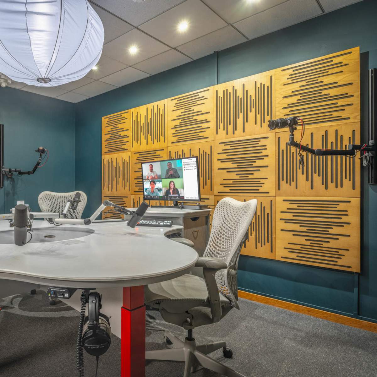 A modern radio studio featuring a round table, adjustable microphones, soundproofing panels on the walls, and a workstation with a monitor.
