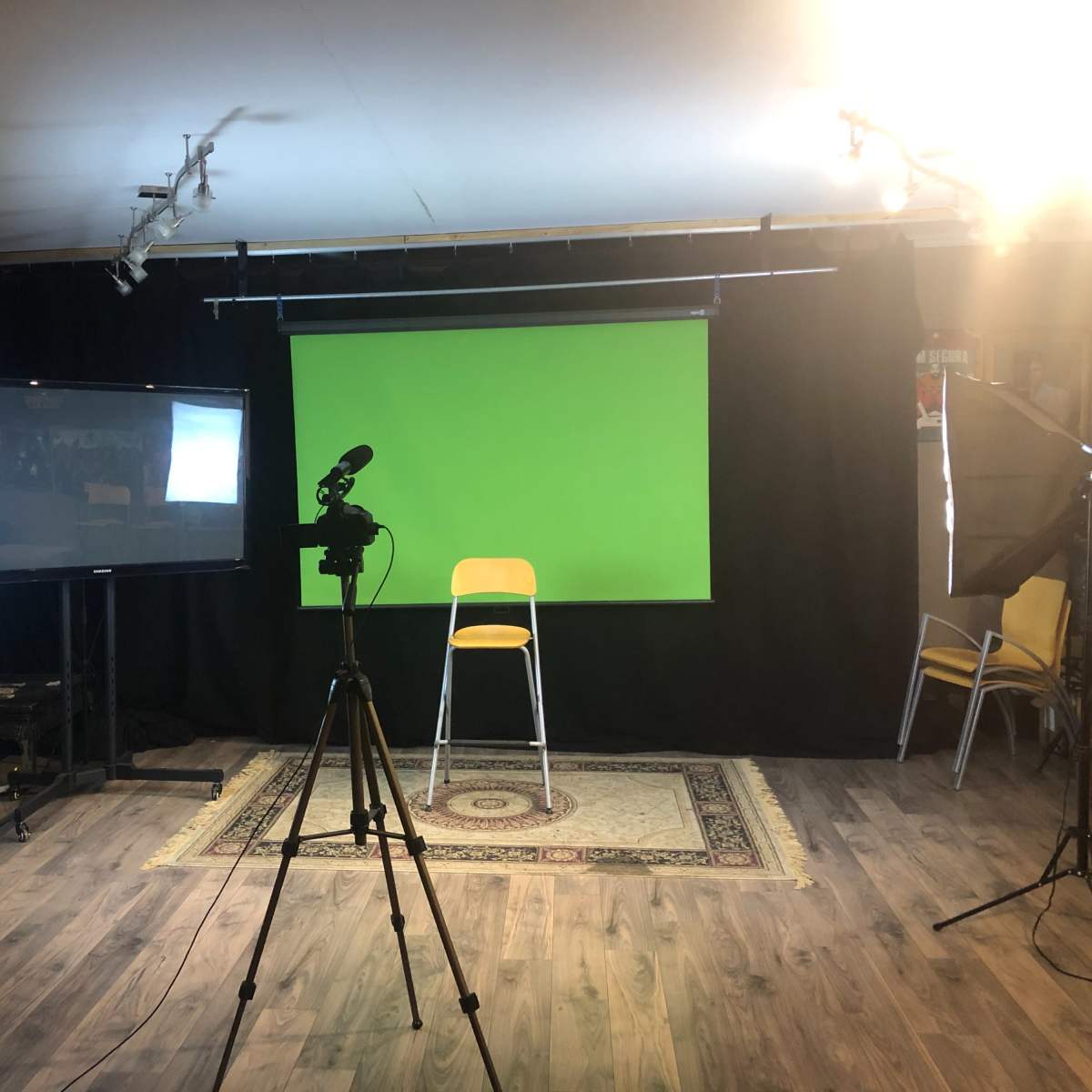 A small studio setup featuring a green screen, a camera on a tripod, a TV monitor, and lighting equipment, all arranged around a wooden floor with a patterned rug.