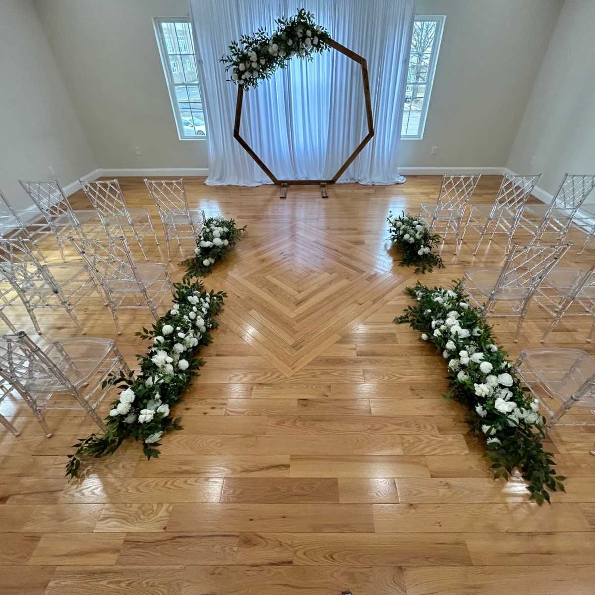 A wedding ceremony setup featuring clear chairs arranged around a floral-accented aisle, leading to a hexagonal wooden arch draped with fabric.