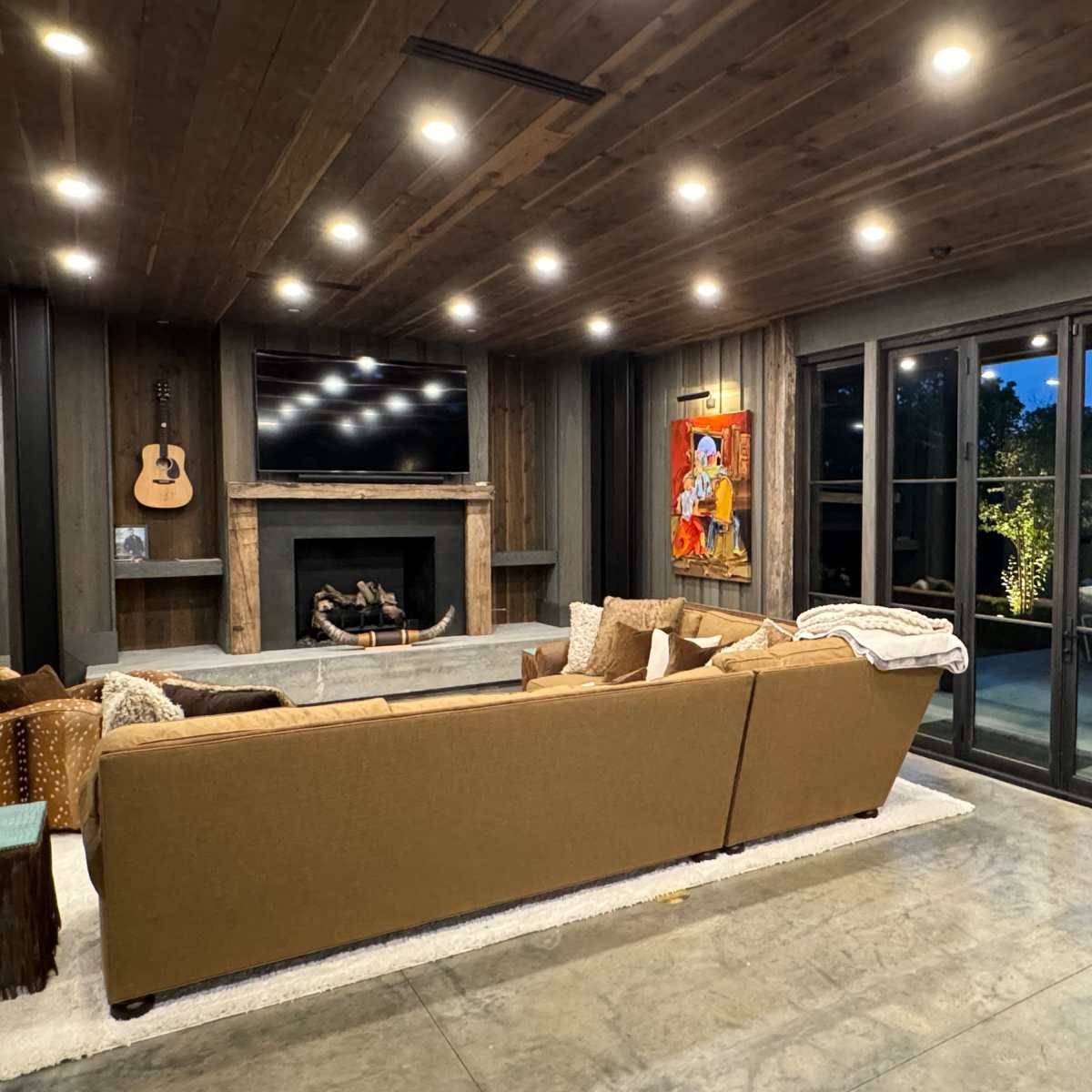 A modern living room with a brown sectional sofa, a fireplace with a television above it, large windows, and a wooden ceiling.