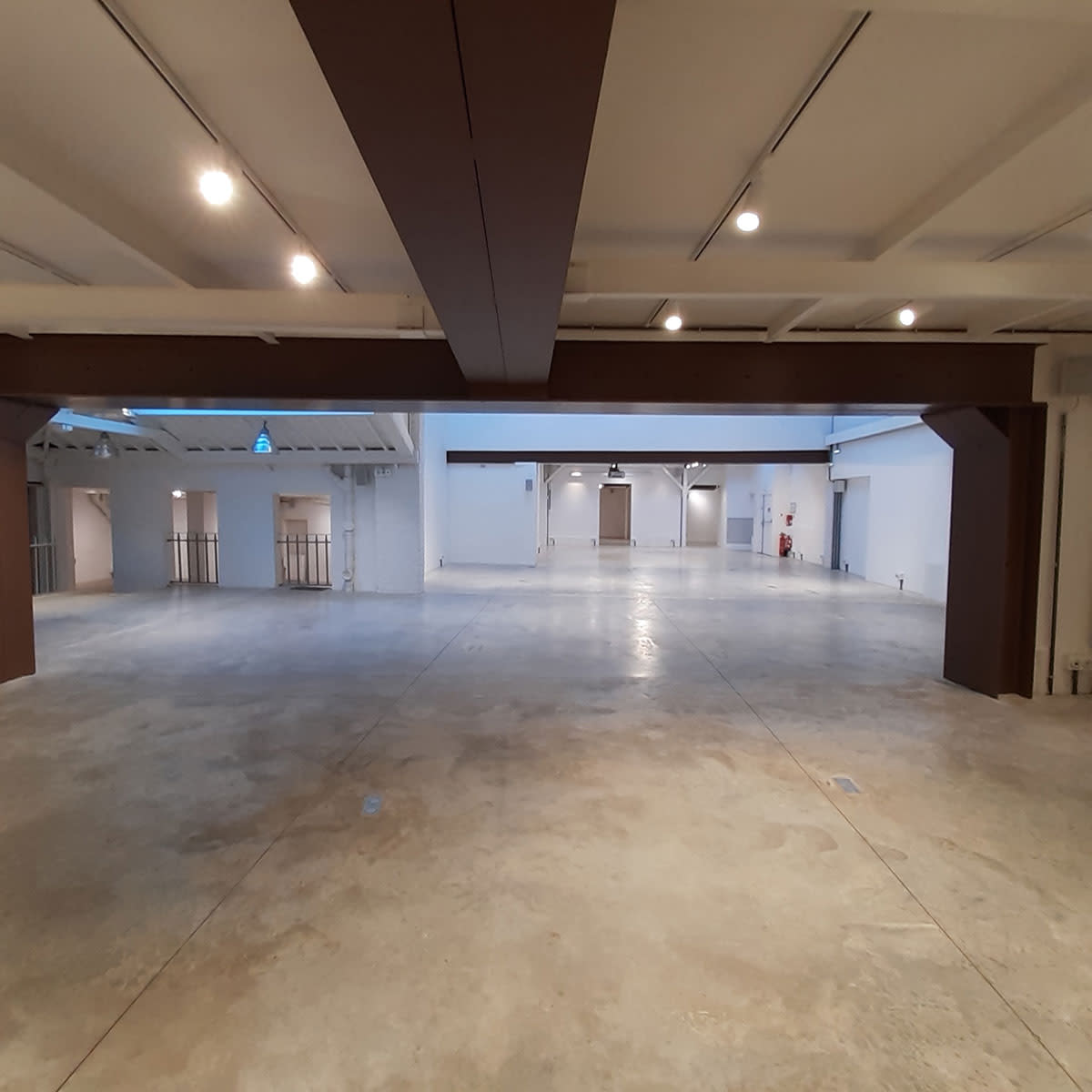 A spacious, empty interior with a polished concrete floor, illuminated by overhead lighting and featuring white walls and wooden beams.