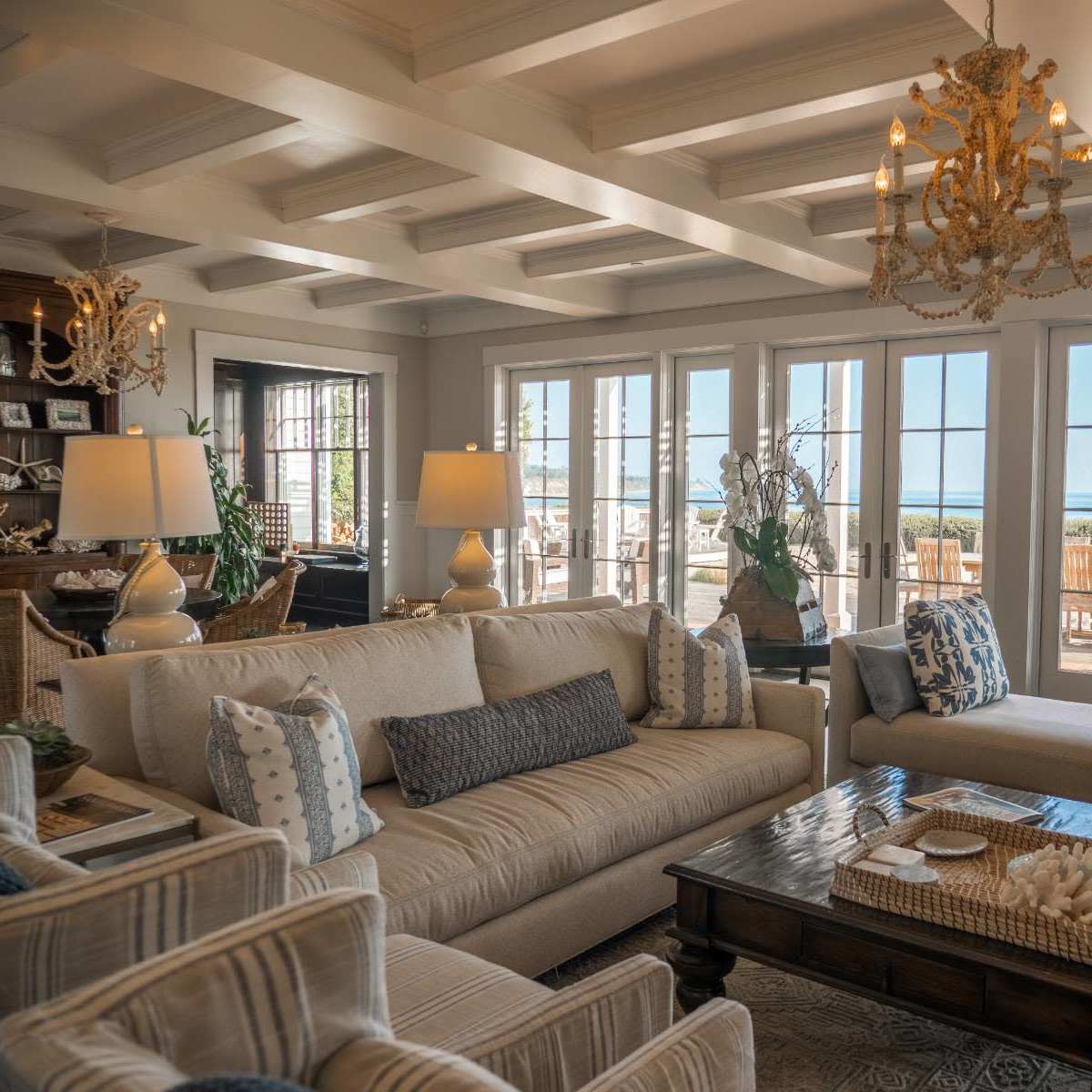 A bright living room with a large beige sofa, patterned armchairs, and a wooden coffee table, featuring large windows that open to a view of an outdoor dining area and the water.