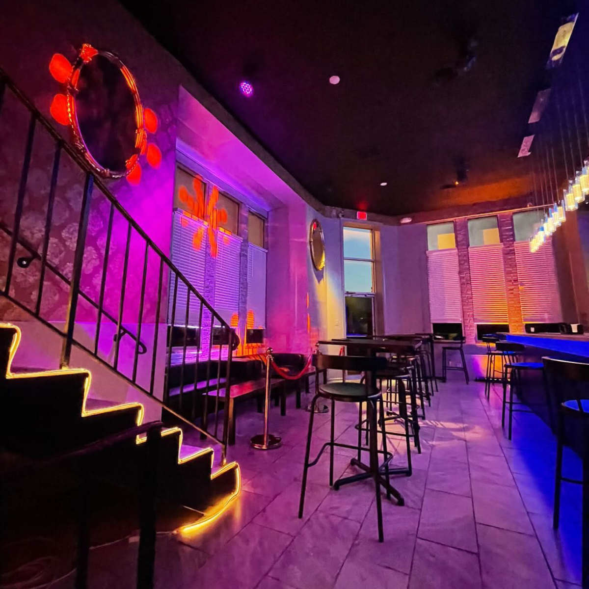 A dimly lit bar area with colorful lighting, staircase, and rows of high tables and chairs.