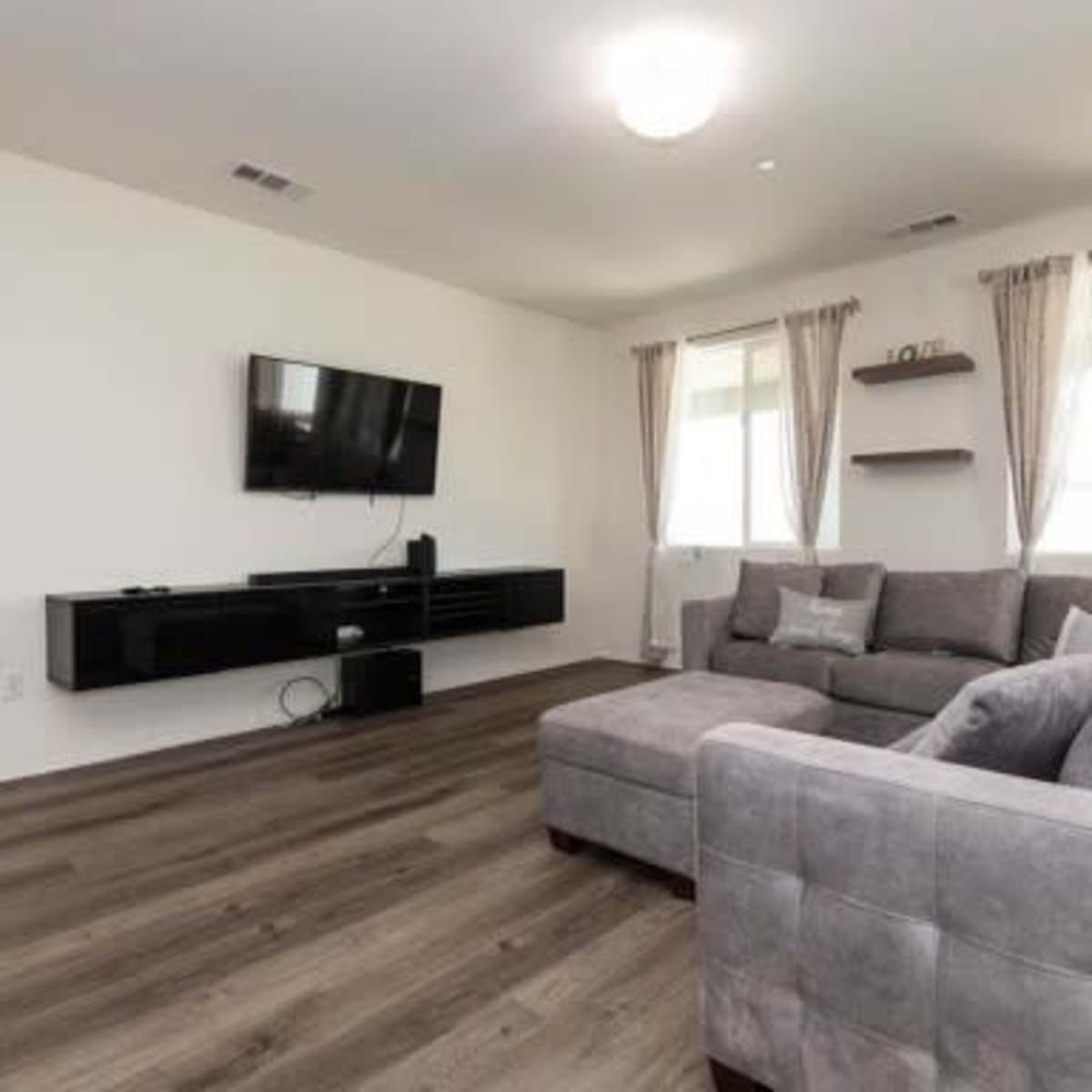 A modern living room with a gray sectional sofa, a wall-mounted television, and large windows dressed with light curtains.