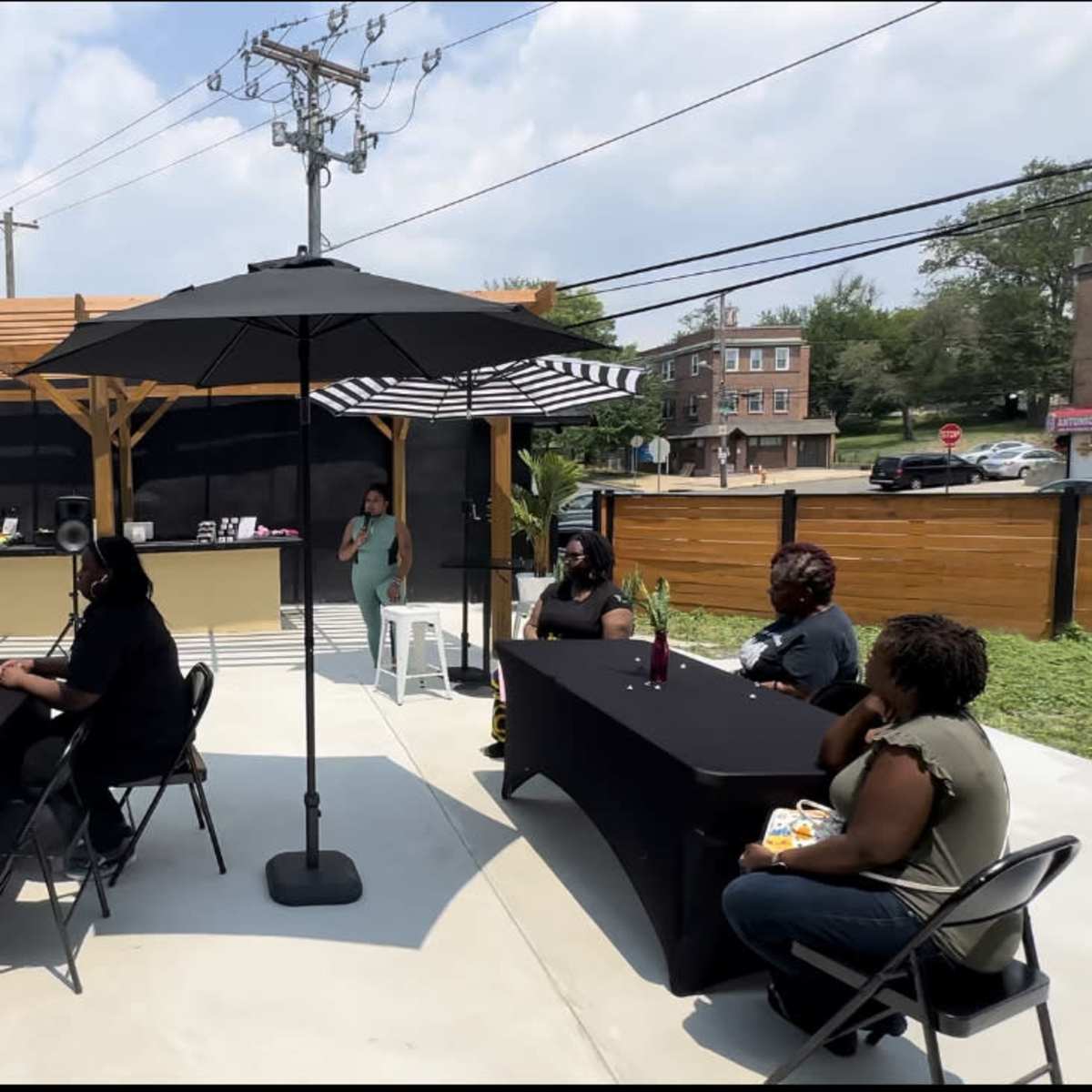 A group of people sits at tables under large umbrellas in an outdoor gathering space, while a person stands at the front speaking.