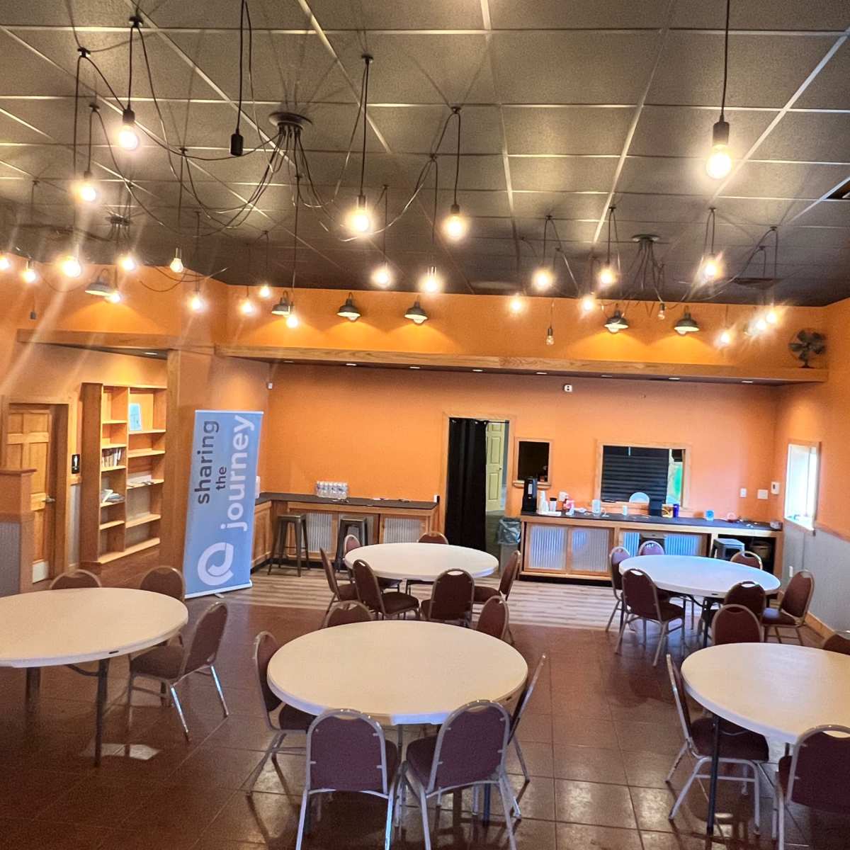 A spacious community room is arranged with several round tables under hanging lights, featuring a banner for "Sharing Journey" and a serving area in the background.