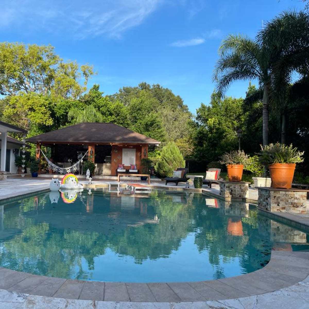 A residential backyard with a large swimming pool surrounded by stone borders, a gazebo, potted plants, and sun loungers.