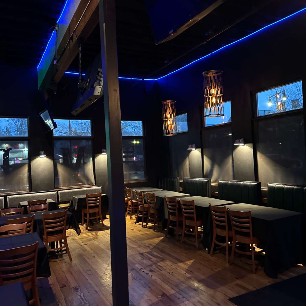 An empty restaurant interior with wooden flooring, dark walls, and several tables arranged with black tablecloths under dim lighting.