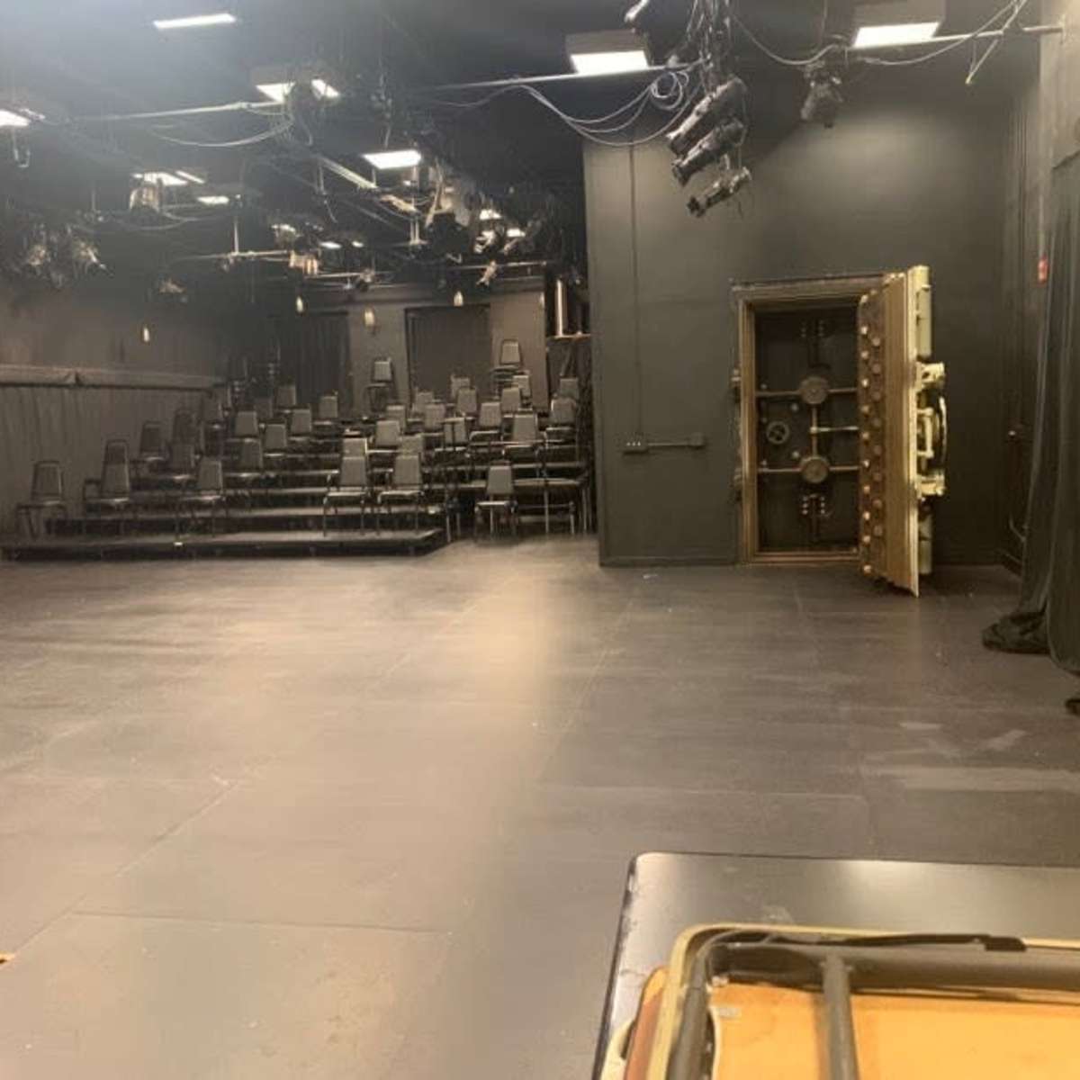 A black-box theater with rows of stacked chairs, a safe door on the right, and dark curtains in the background.