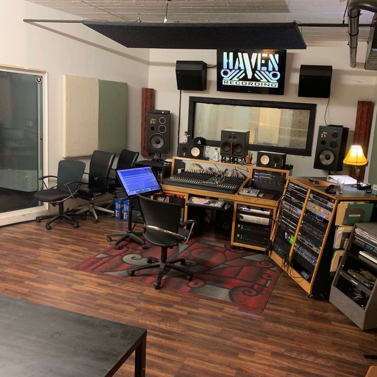 A recording studio with a console, speakers, a computer setup, and several chairs arranged around a central carpeted area.