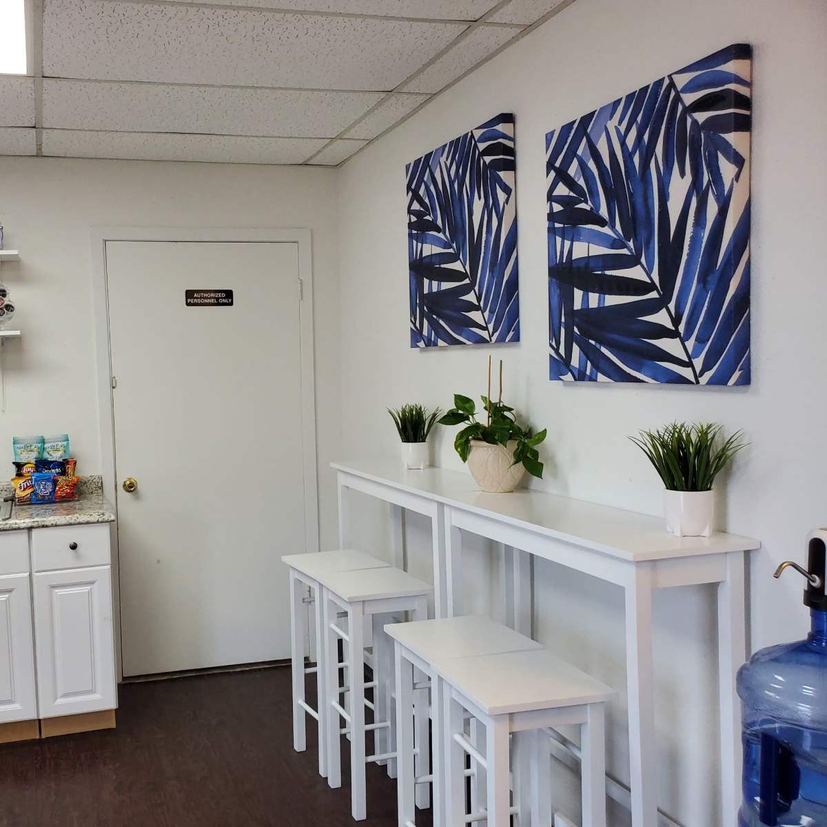A kitchenette area with a countertop, a sink, two paintings of blue leaves on the wall, a small table with three stools, and a water dispenser.