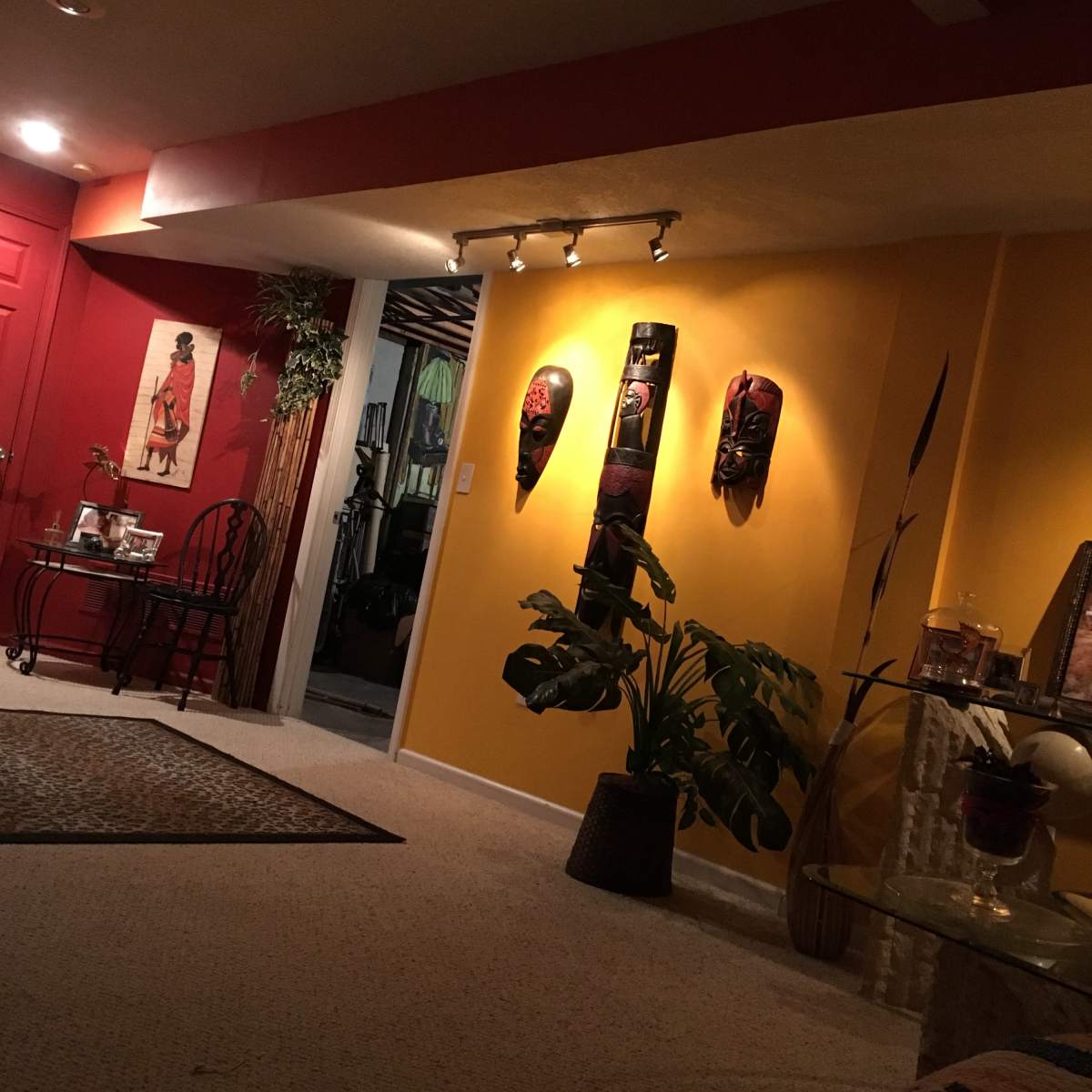 A warmly lit living space with a yellow wall adorned with three decorative masks, a potted plant in the foreground, and a red wall with a door in the background.