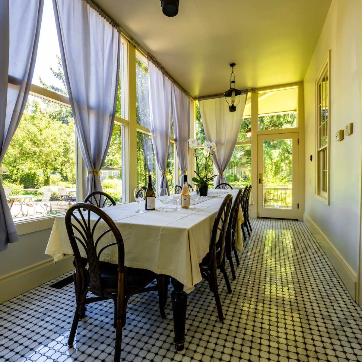 A long table is set with bottles and a tablecloth in a sunlit room with large windows and patterned tile flooring.