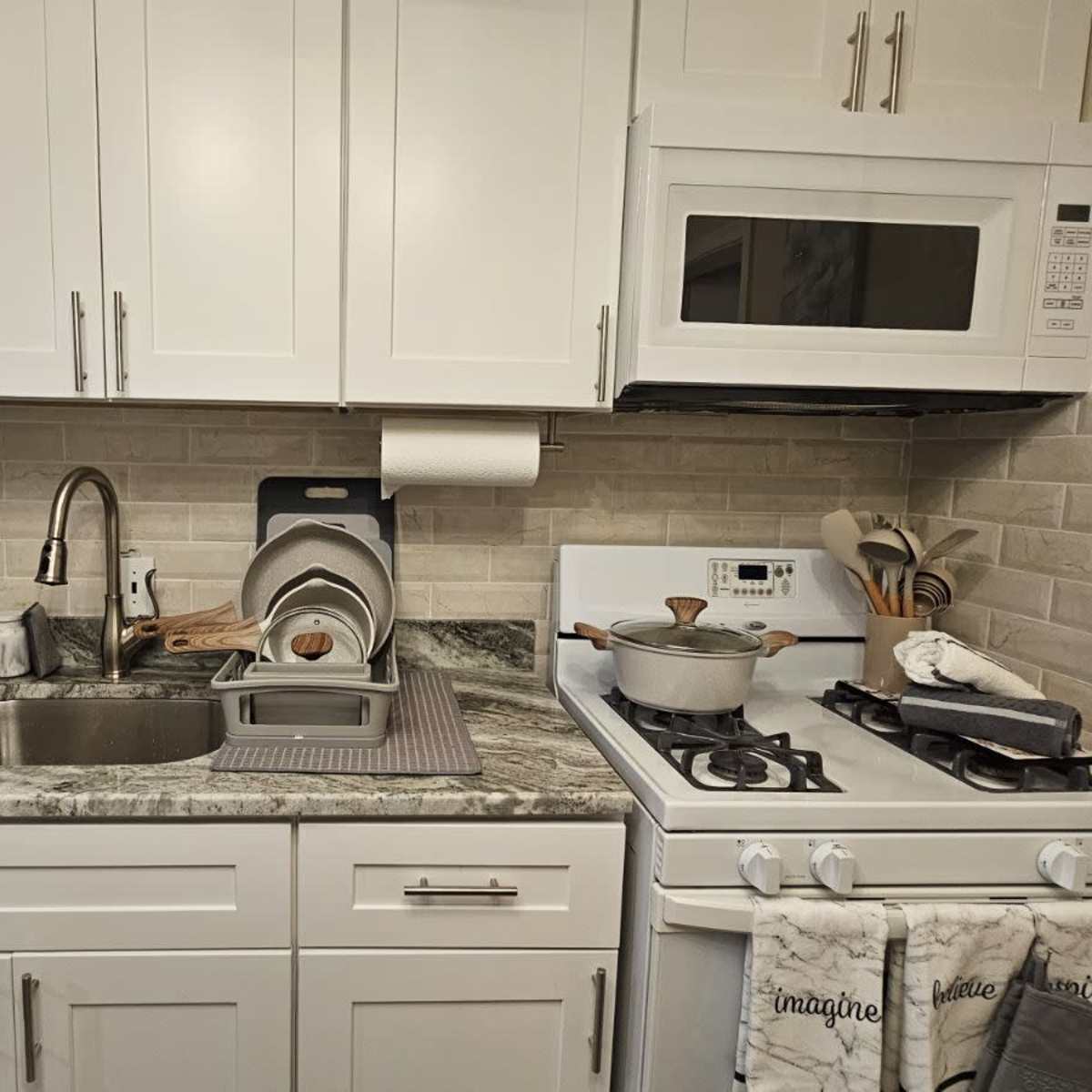 A modern kitchen with white cabinets, a sink, a stovetop, and a microwave, all featuring a stone countertop and gray tile backsplash.