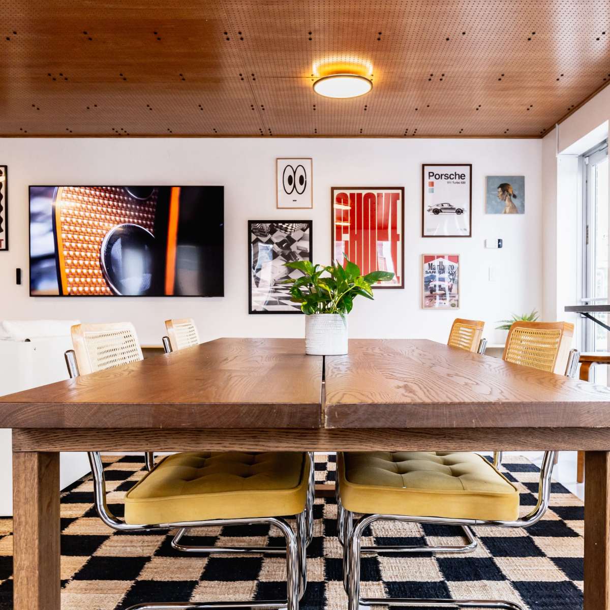 A wooden dining table with yellow chairs is set in a room featuring a wall of framed artwork and a television.